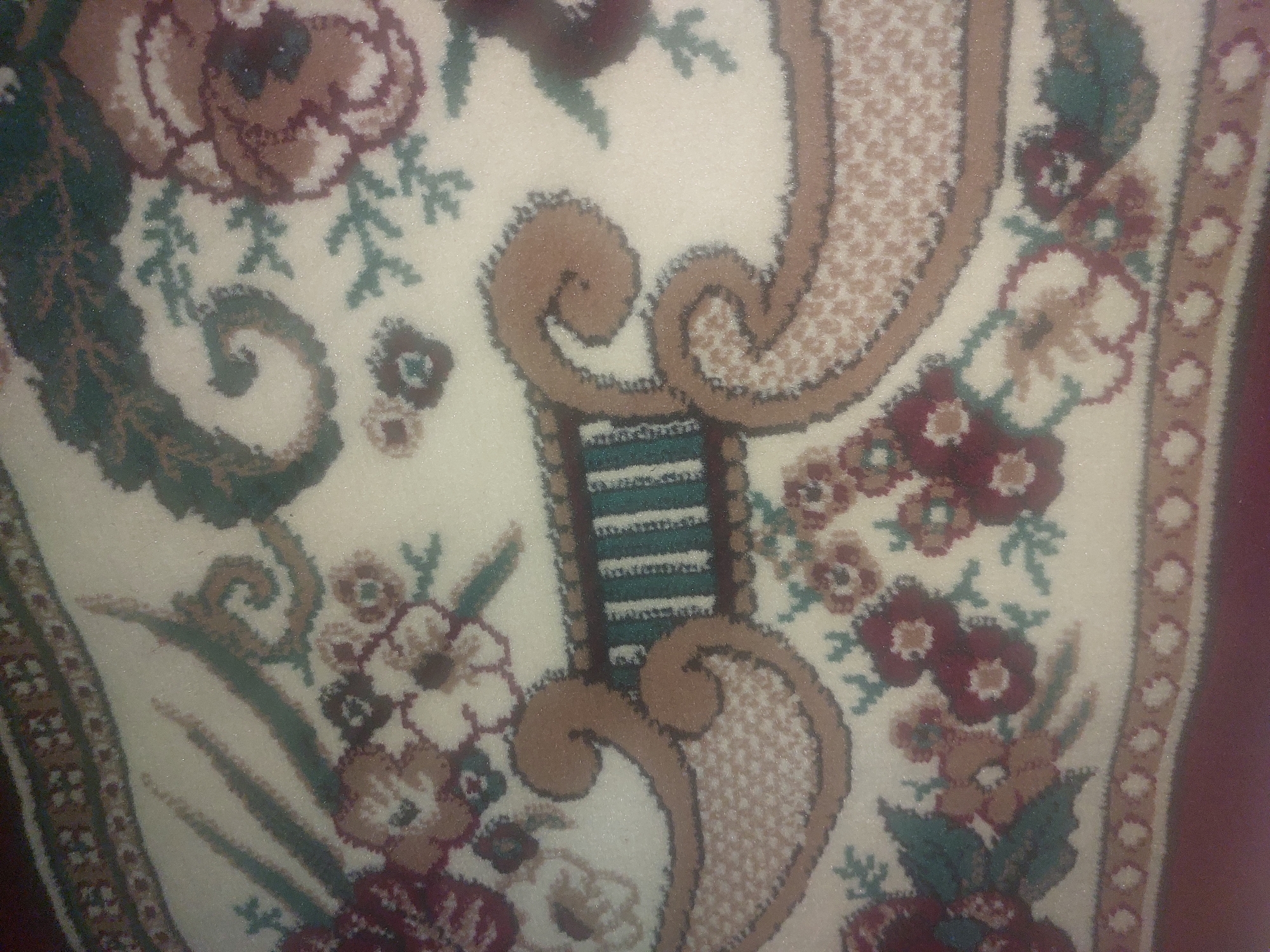 This is a close-up, high-angle, slightly blurry photograph of a portion of an ornate rug. The rug has a cream-colored background with a repeating pattern of stylized floral elements and scrolling motifs in shades of burgundy, green, and tan. The rug appears to be lying flat on a surface, possibly a floor. There are no people or other discernible objects visible in the image. The image provides no information about the location in the city of Ması, Kyrgyzstan, or any contextual details beyond the rug itself.