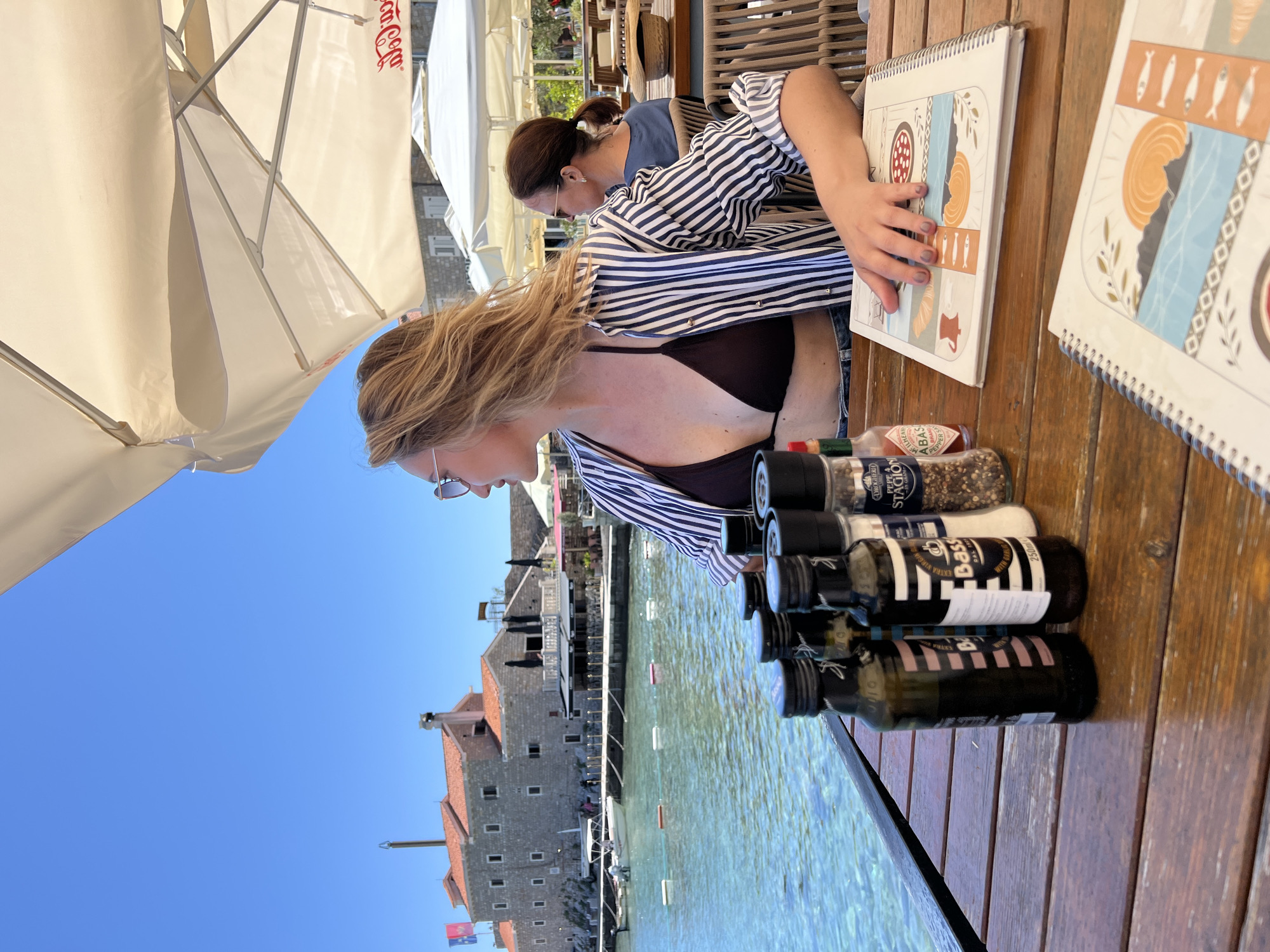 This image depicts an outdoor dining scene at a waterfront restaurant in Petrovac centar, Montenegro, on a bright, sunny day. Two women are seated at a wooden table, engaged in selecting food from menus. The woman in the foreground has long blonde hair, wears sunglasses, a dark bikini top, and a blue and white striped shirt worn open. The second woman, seated further back, has dark hair tied up and is also wearing a striped shirt, with her hand resting on a menu.
The wooden table holds several condiments: bottles of olive oil (one labeled "Bass" with "EXTRA VIRGIN OIL" and "250ml"), salt and pepper grinders (one visible with "DROGHERIA" and "PEPE STAGION"), and a small red bottle of "TABASCO BRAND PEPPER SAUCE" by "ILHENY COMPANY". Two white spiral-bound menus, adorned with colorful illustrations of fish, food items, and abstract patterns, are laid out.
The background showcases a vibrant coastal setting with a clear blue sky. Overhead, large white patio umbrellas provide shade, one prominently displaying a "Coca-Cola" logo. Beyond the dining area, tranquil, clear blue water stretches out, dotted with a few boats. On the far side of the water, historical stone buildings with distinctive red-tiled roofs line the shore, indicating the charming architecture of the region. The overall atmosphere appears relaxed and pleasant under the bright sunshine.