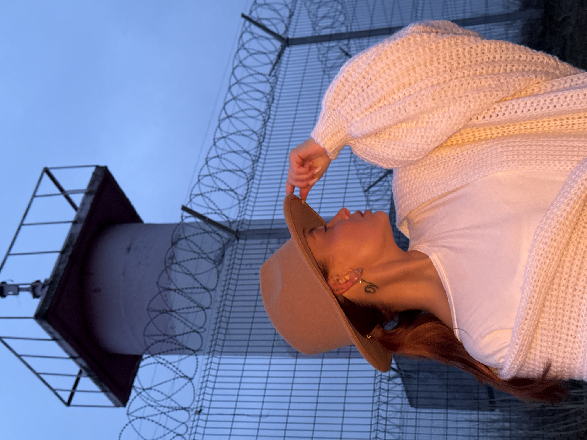 This image captures a young woman in profile, positioned on the right side of the frame, looking upwards with a contemplative expression. She wears a white t-shirt beneath a chunky, off-white knit cardigan with voluminous sleeves. A light brown fedora-style hat is on her head, which she delicately touches with her right hand. Her reddish-brown hair flows down her back, and a small, stylized tattoo is visible on her neck near her ear, which also sports an earring.
The background features a deep blue sky, suggesting either dusk or dawn. On the left, a tall, dark tower or mast structure stands, topped with a square platform, resembling a surveillance or technical installation. An extensive fence, constructed from wire mesh and topped with prominent coils of razor wire, dominates the middle ground, extending across the frame and creating a sense of enclosure around the tower. The fence angles diagonally, emphasizing the secure or restricted nature of the location. The soft, ambient light illuminates the woman's face and neck with a gentle, warm glow. The scene is set outdoors in what appears to be an industrial or controlled area, contextually located in Chaikovsky, Russia. There is no visible text in the image.