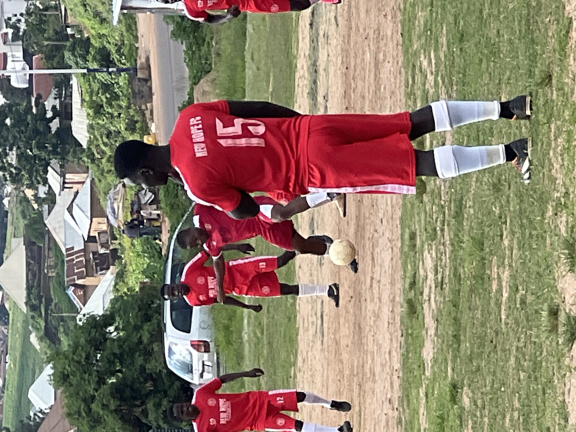 The image captures a casual soccer match in Jos, Nigeria. Four young men, all wearing matching red jerseys and shorts, are actively engaged in the game. Three of the players are visible from the waist up, while the fourth is shown from the knees down, focusing on their footwear. One player in the foreground, facing away from the camera, has the number "15" prominently displayed on the back of his jersey, along with the text "NEW HOPE FC". Another player, to the left, wears the number "12". A soccer ball is visible on the dusty ground, being kicked by one of the players. The background reveals a mix of residential buildings and lush greenery, characteristic of the city's landscape. A white vehicle is parked to the left, adding a touch of everyday life to the scene. The overall atmosphere suggests an informal sporting event taking place outdoors.