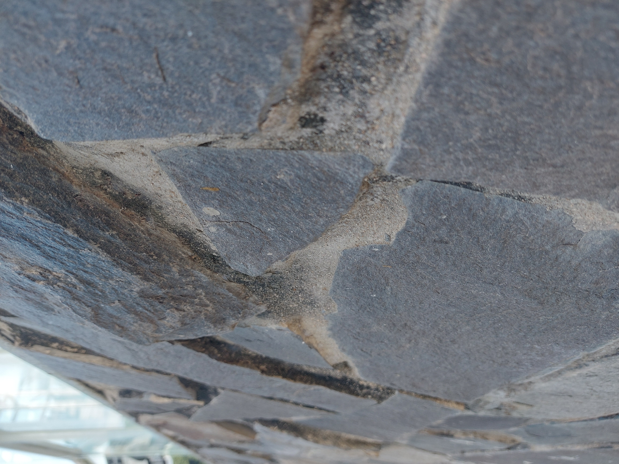 The image is a close-up, side-view, outdoor photograph of a stone wall constructed with irregularly shaped, dark gray stones of varying sizes. The stones are mortared together with a light-colored cement. The wall extends diagonally from the bottom left to the top right of the frame. The texture of the stones is rough and appears to have natural variations in color and surface. The lighting suggests it is daytime, with some shadows present between the stones. In the blurred background on the left, a portion of a modern building with glass windows and possibly some greenery is visible, indicating an urban environment. The composition is focused on the details of the stonework, with a shallow depth of field blurring the background. The location is specified as Nha Trang Ward, Vietnam.