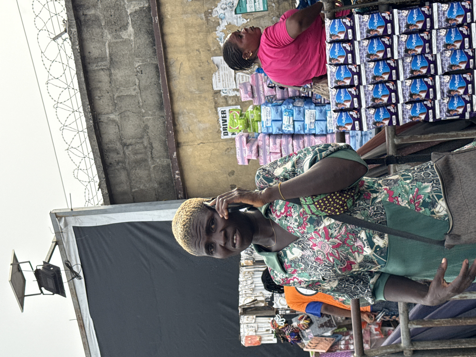 This image captures a bustling street market scene, likely in Lagos, Nigeria, under bright daylight.
In the foreground, a woman with short, bleached blonde hair and dark skin is visible, holding a dark-colored smartphone to her right ear, appearing to be on a call. She wears a vibrant, long-sleeved shirt with a green, pink, and white floral pattern, layered over a solid green garment, and a black bag strap crosses her body. She looks directly towards the viewer with a calm expression, showing a slight smile.
Behind her, a merchandise stall is prominent, displaying numerous packs of sanitary pads. Many of these are branded "Softcare," with some featuring blue packaging and others pink. The packs are arranged neatly, some hanging vertically, others stacked.
Further in the background, another woman with braided dark hair and wearing a bright pink t-shirt is seen looking upwards, possibly tending to her stall or observing something above. Her stall also features stacks of sanitary pads. Behind her, a weathered yellow wall is visible, adorned with various signs, including one that clearly reads "DRIVER" and "VACANCY." The wall shows signs of wear and tear, with peeling paper.
To the upper left of the image, barbed wire is visible against a bright, overcast sky, suggesting a security fence or barrier. A black fabric sheet serves as a backdrop, possibly for a stall, and a small light fixture is mounted on a metal frame nearby. The overall setting portrays a typical lively outdoor market environment.