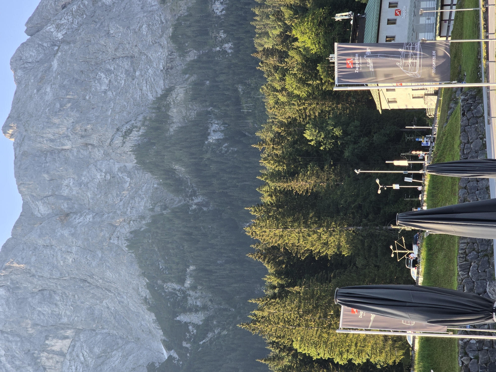 This image depicts a view of the Zugspitze mountain in Grainau, Germany, as seen from a lower elevation. In the foreground, several large, dark gray umbrella covers are closed and standing upright. To the right of the umbrellas, a tall flag with an illustration of a cable car and the text "Zugspitze.de" is displayed. Behind the flag, a light-colored building with a green roof and multiple windows is visible. This building appears to be a visitor center or station, as suggested by the signage that includes "Zugspitze TOP OF GERMANY." A street with several parked cars and a few people is situated in front of the building and the trees. The middle ground is dominated by a dense forest of evergreen trees, which gives way to the imposing, rocky face of the Zugspitze mountain in the background. The sky is clear and blue. The scene is illuminated by sunlight, suggesting it is daytime.