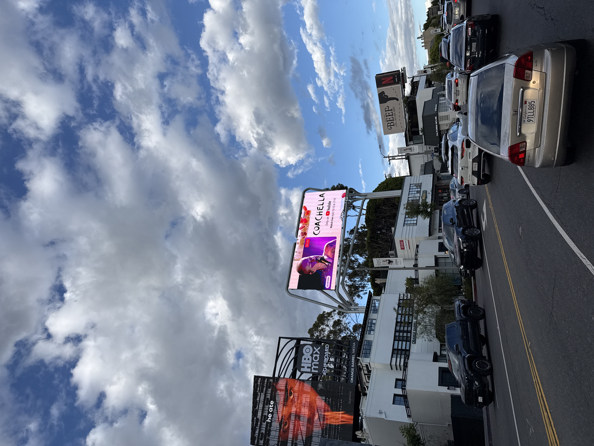 The image captures a street scene in Los Angeles, filled with a mix of billboards and traffic. Dominating the view are several large billboards displaying advertisements for HBO Max's "Euphoria", Coachella, and Netflix's "Beef". The "Euphoria" billboard shows a close-up of a person's face, while the Coachella billboard promotes the live streaming of the music festival on YouTube. The "Beef" billboard shows an animal with an axe. The sky above is a mix of blue and cumulus clouds. A row of cars is lined up on the street, including sedans and trucks. California license plates are visible on some of the vehicles. The road is asphalt. Buildings are visible behind the billboards and the cars. The image gives the impression of a bustling urban environment with heavy traffic and a concentration of advertising.