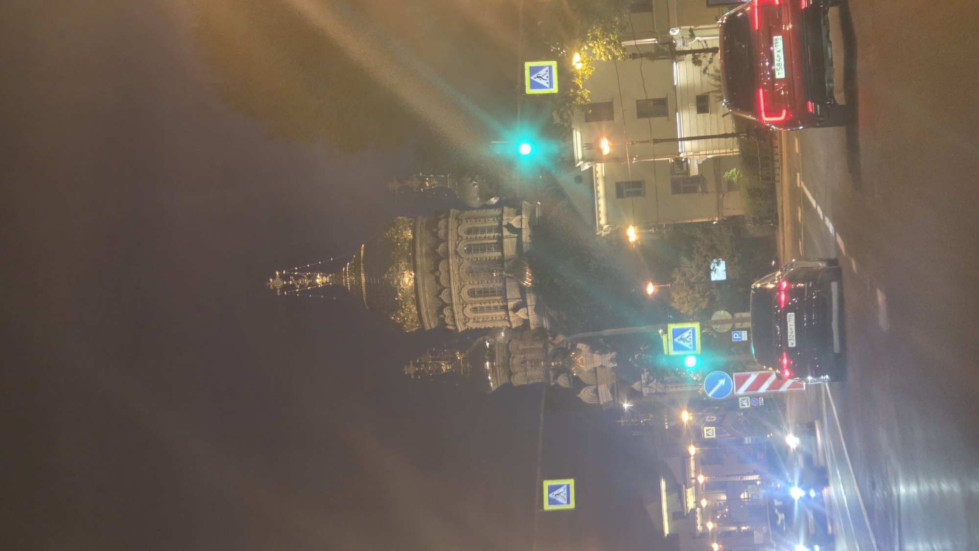 The image depicts a night scene in Unknown, Unknown, showing a street with two cars driving on it. On the left side of the street is a large, ornately decorated building with golden domes, possibly a church or cathedral. There are streetlights illuminating the buildings and street. Traffic lights are visible, indicating that the cars are at an intersection. Pedestrian crossing signs are also present. The sky is dark, suggesting it is nighttime.