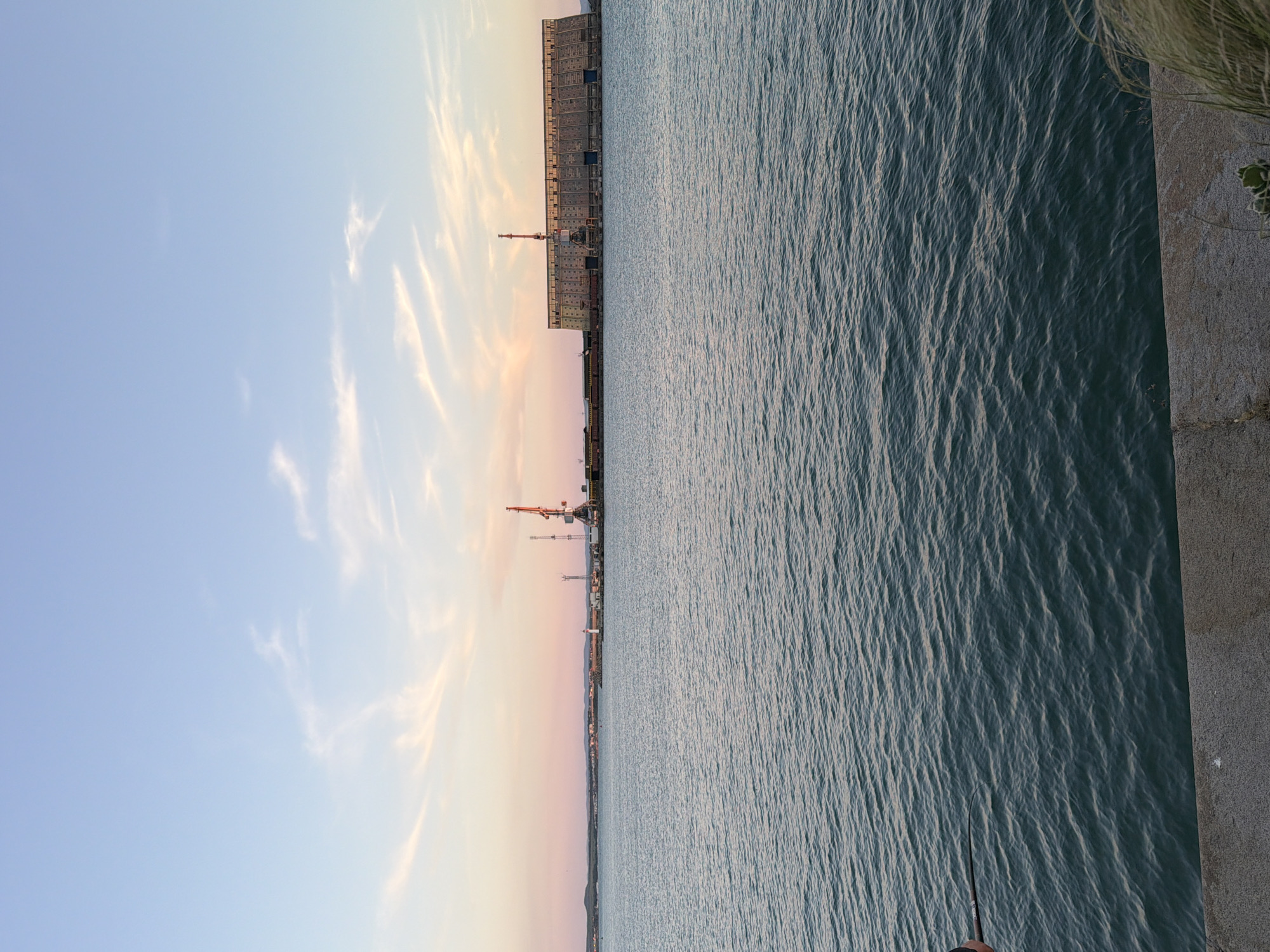 The image depicts a wide shot of the sea at sunset, with the city of Burgas, Bulgaria, visible in the distance. The sky is a blend of soft blues and pinks, with wispy clouds. The sea is calm, with gentle ripples reflecting the sky's colors. In the mid-ground, there's a large industrial structure, possibly a shipyard or factory, with cranes and other equipment. A lighthouse is also visible on the shore. The foreground shows a concrete embankment with some dry grass. The lighting suggests it is late afternoon or early evening.