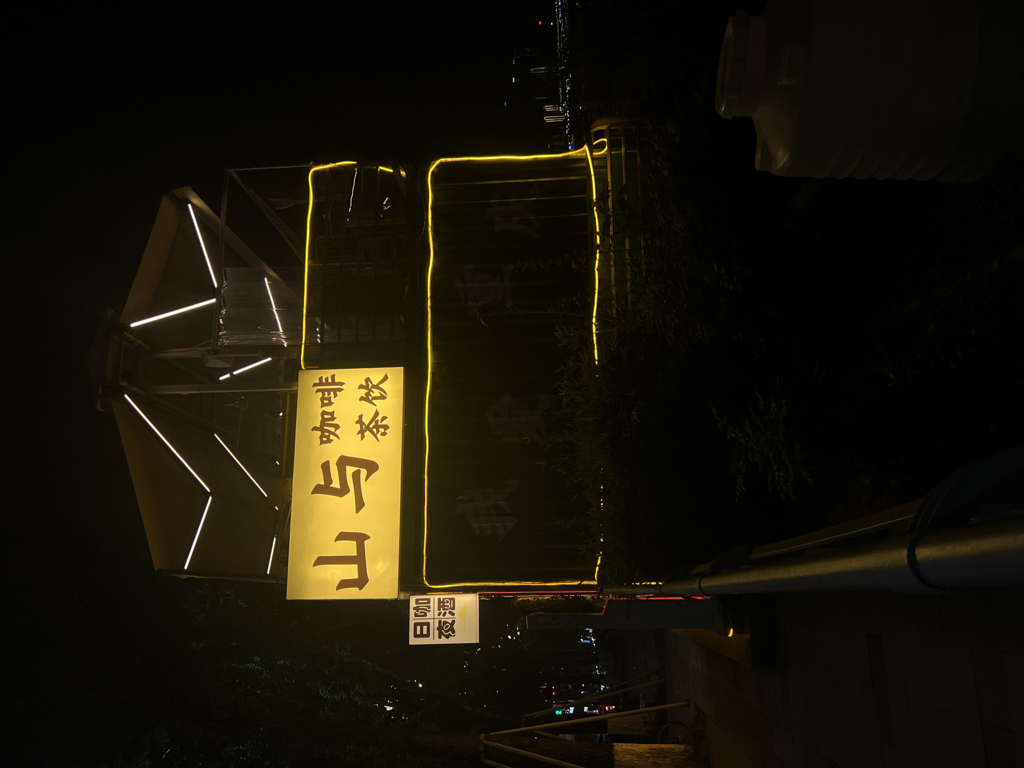 The image shows a night scene in Nan'an District, China, illuminated by artificial lights. A prominent yellow sign with Chinese characters reads "山与咖啡茶饮," which translates to "Mountain and Coffee Tea Drinks." Below this, another smaller sign displays "日咖夜酒," meaning "Coffee by day, drinks by night." The sign is attached to a structure with a modern design, outlined with bright yellow neon lights. The structure appears to be an outdoor seating area or an establishment's facade. In the background, city lights are visible in the distance, suggesting an urban environment. The foreground features a sidewalk and a metal railing, with some vegetation present. A large, ribbed plastic container is visible on the right side of the frame. The overall impression is of a commercial establishment at night, likely a cafe or bar.