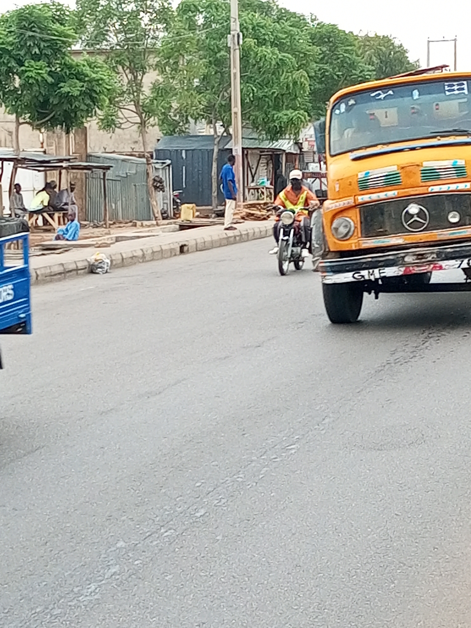 The image shows a street scene in Gombe, Nigeria. A yellow Mercedes-Benz truck is on the right side of the road, facing the camera. A motorcyclist wearing a white cap and an orange vest is riding past the truck. Several people are gathered under a shelter on the left side of the road, and another person is standing near a blue container. A small blue vehicle is partially visible on the left. The road surface is asphalt. Trees and buildings are visible in the background.