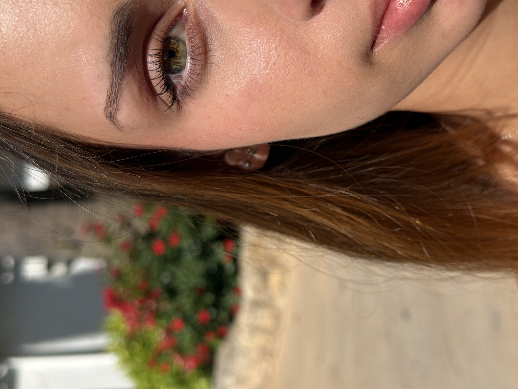 A close-up, rotated portrait shows a portion of a woman's face, including her eye, cheek, upper lip, and hair. Her eye has long lashes and a mixture of green and brown colors in the iris. Her skin is fair, with a slightly pinkish tint. Her lips appear to have a glossy finish. She has brown hair.
In the blurred background, there is greenery with red flowers. A building structure is also visible. The location is in Bunyola, Spain, as stated in the prompt.