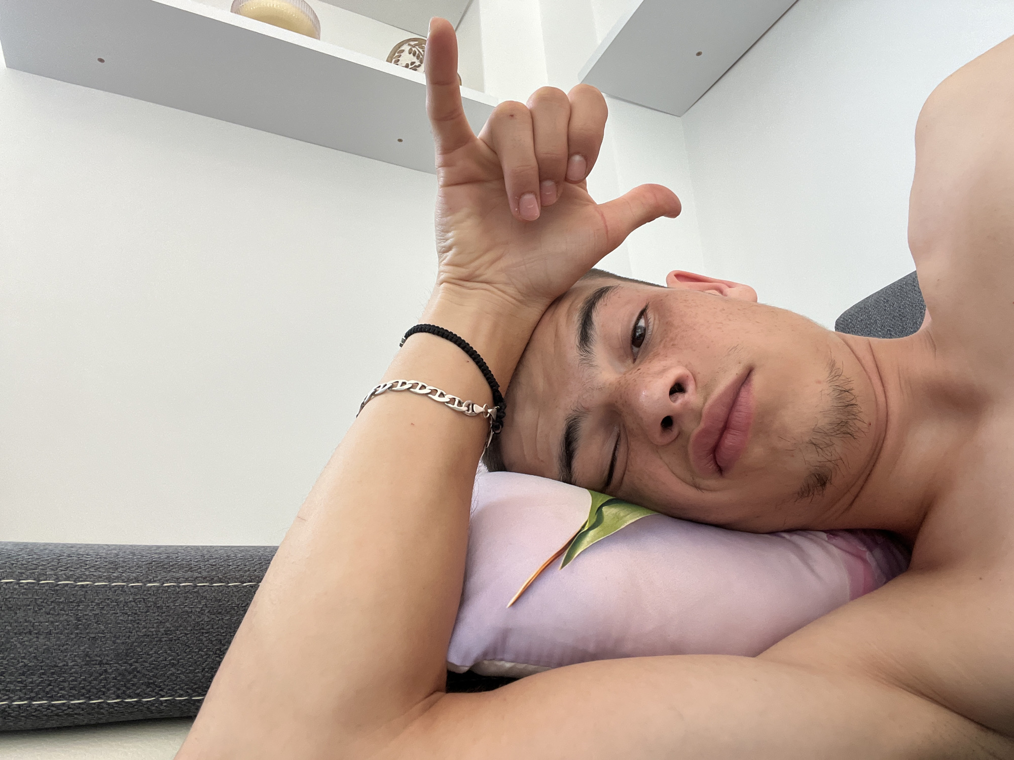 A young man is lying on his side on a pillow, resting his head on it. He is looking at the camera with a relaxed expression. His right arm is bent at the elbow, with his hand raised to his forehead, fingers curled in a gesture. He is wearing a silver bracelet on his left wrist. The background is a white wall with a shelf above. The scene appears to be indoors, likely in a bedroom, given the presence of a pillow and the relaxed posture of the subject. The overall impression is one of informality and personal intimacy. There is no identifiable information that places this image specifically in Karavelovo, Bulgaria; it is a generic interior setting.