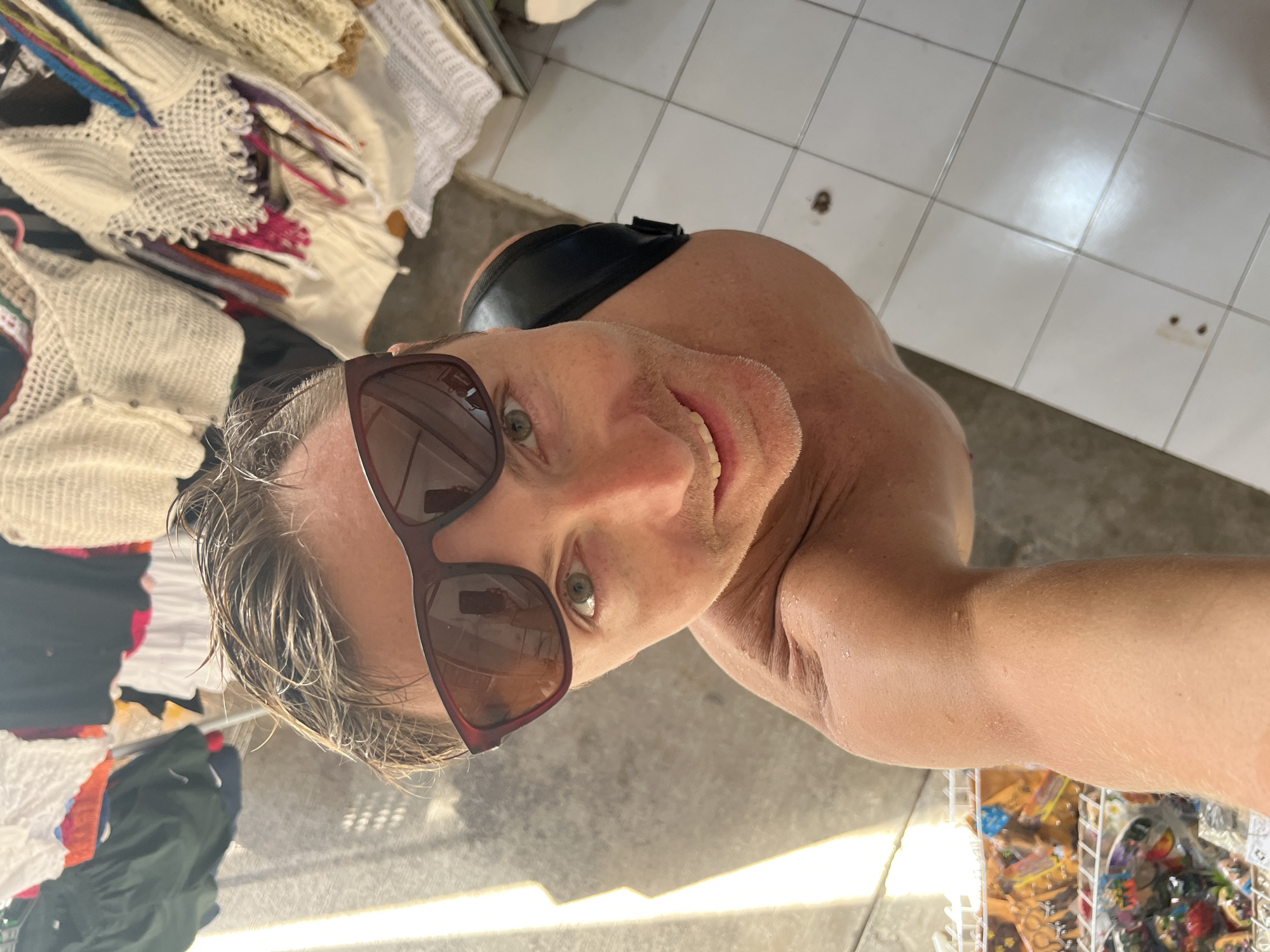 The image is a close-up selfie of a fair-skinned man with short, wet blonde hair and brown sunglasses. He is smiling and looking into the camera. He appears to be shirtless, with a black strap visible across his upper back. The background shows elements of an indoor market setting. To the left are racks of white crochet clothing and other garments. To the right is a tiled floor and a wire basket filled with packaged goods, likely toys or souvenirs. The location is indicated as Kuta Selatan, Indonesia. The overall impression is that the man is a tourist engaging in commerce at a local market in Kuta Selatan.