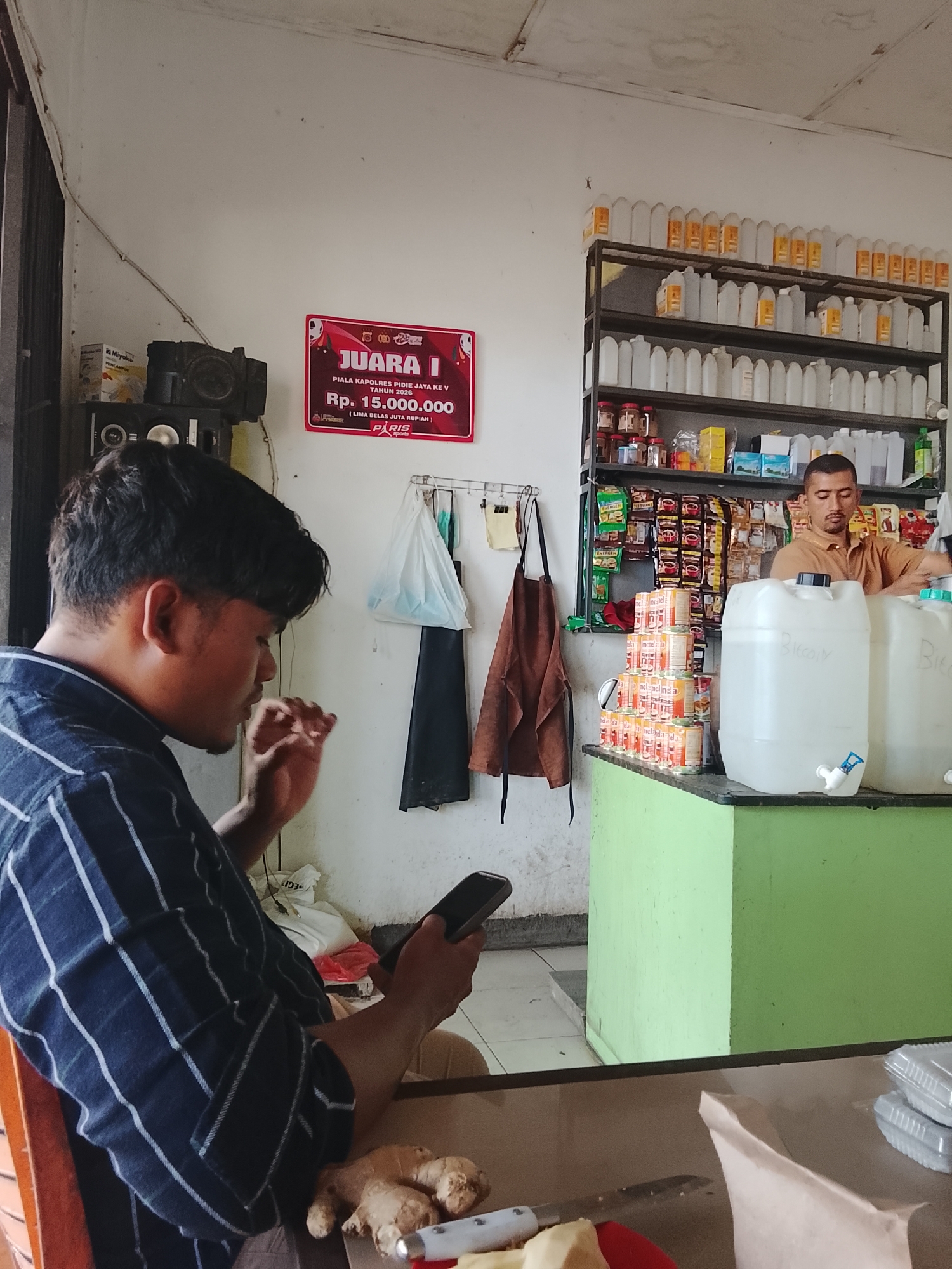 The image captures an indoor scene in what appears to be a small shop or store, possibly in Meunasah Pupu, Indonesia, given the information provided. In the foreground, a young man with dark hair is seated, facing to the right and looking down at a mobile phone he is holding. He is wearing a dark blue and white striped shirt. To his left, on a table or counter, are several pieces of ginger and a knife with a red and white handle.

Behind the young man, the environment includes shelves stocked with various items. On the left side, there's a speaker and a framed award or poster that reads "JUARA I PIALA KAPOLRES PIDIE JAYA KE V TAHUN 2026 Rp. 15.000.000". Further back on the left, there are several plastic bags hanging.

The right side of the image features a man with short dark hair and a light brown shirt standing behind a green counter. He is interacting with something behind the counter. In front of him are two large, clear plastic containers with spouts. The shelves behind him are filled with bottles and packaged goods, including what look like canned drinks and jars. A stack of orange-colored cans is visible on the counter. The walls are painted white, and the ceiling appears to be made of fiberboard. The overall impression is that of a modest retail space selling a variety of products.