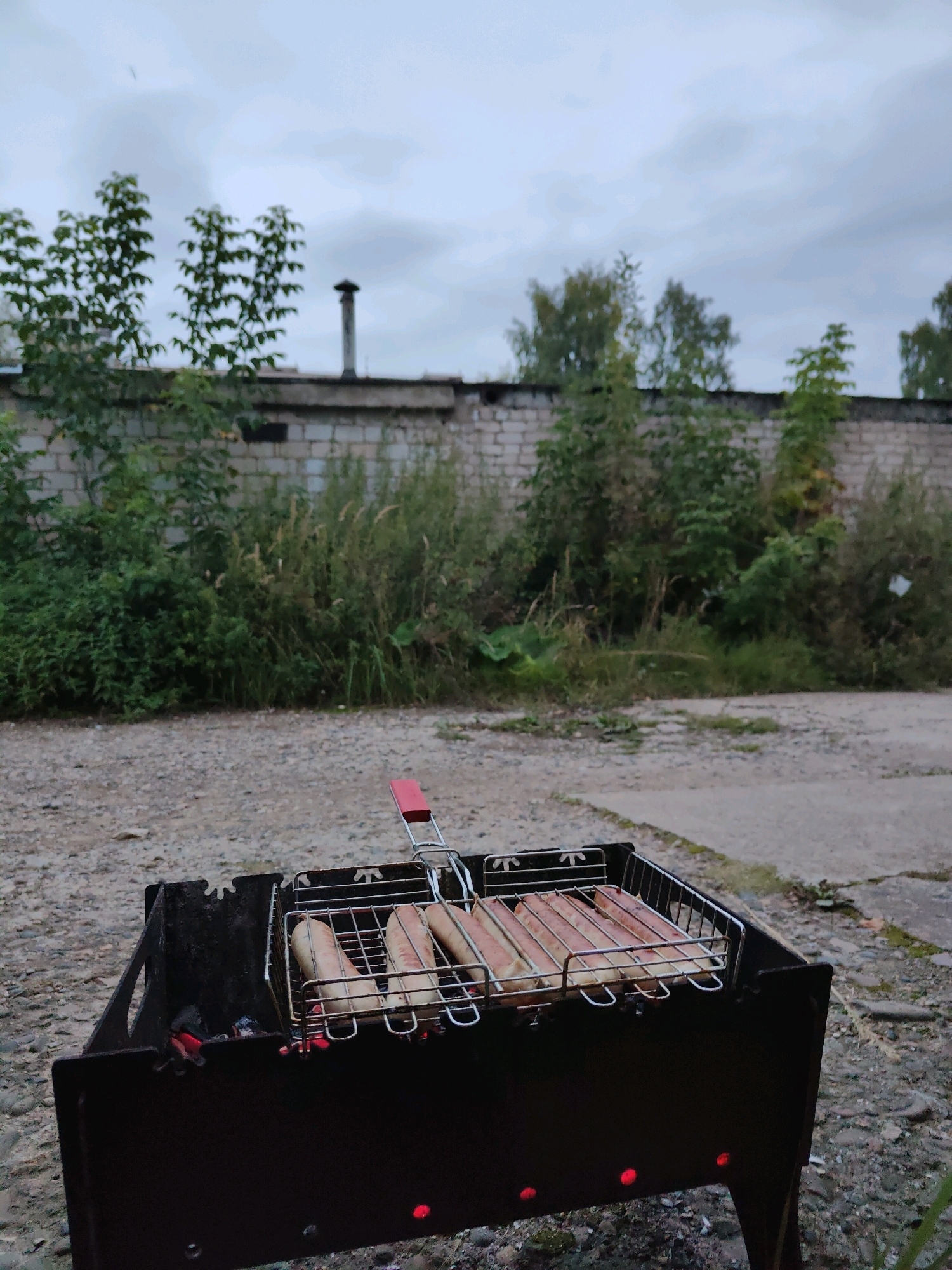 A charcoal grill with sausages cooking sits in the foreground. The grill is made of black metal and is placed on a gravel surface. Several sausages are arranged in a grill basket over glowing red embers. In the background, a low brick wall separates the area from a patch of overgrown vegetation and trees. A tall, narrow chimney rises behind the wall. The sky is overcast and gray. The scene appears to be set in an outdoor industrial or semi-industrial area, possibly on the outskirts of Chaikovsky, Russia, given the overall aesthetic and vegetation. No people are visible in the image.