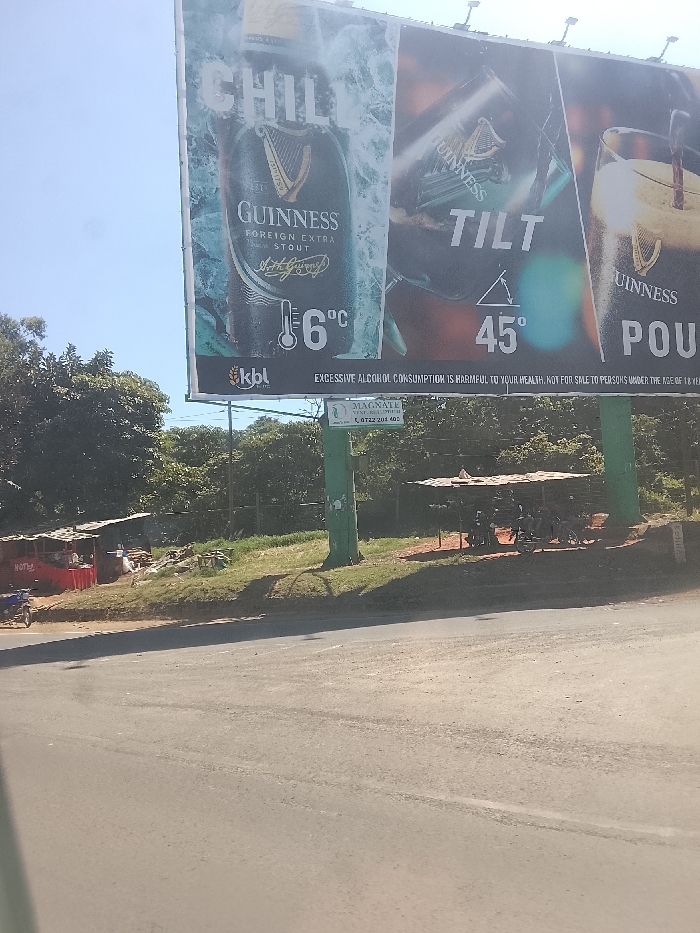 This image depicts a roadside scene dominated by a large billboard advertising Guinness beer. The billboard is divided into panels: the left side displays a chilled can of "GUINNESS FOREIGN EXTRA STOUT" with "CHILL" and "6°C" indicating optimal serving temperature, alongside the "kbl" logo. The right side illustrates the pouring method, with a tilted glass and text "TILT 45°", leading to a full glass with "POUR" and the Guinness logo. Below the main advertisement, a prominent health warning states: "EXCESSIVE ALCOHOL CONSUMPTION IS HARMFUL TO YOUR HEALTH. NOT FOR SALE TO PERSONS UNDER THE AGE OF 18 YEARS."
The billboard is supported by two green pillars, one of which features a smaller sign for "MAGNATE VENTURES LTD" and a phone number (0722 204 400). In the foreground, a paved road stretches across the bottom of the frame. To the left of the road, a small, makeshift stall with a red canopy and the partial text "HOTL" is visible, with a motorbike parked beside it. To the right, beneath a rudimentary shelter constructed from corrugated iron, several people are gathered, possibly sitting or resting.
The background is filled with dense green trees and foliage, suggesting a lush, natural environment. The bright blue sky overhead indicates a sunny daytime. Clear shadows are cast on the ground by the billboard supports and the small structures. The scene portrays a typical roadside in a semi-urban or rural setting, consistent with Kiambu Township ward, Kenya, featuring both commercial advertising and local commerce.