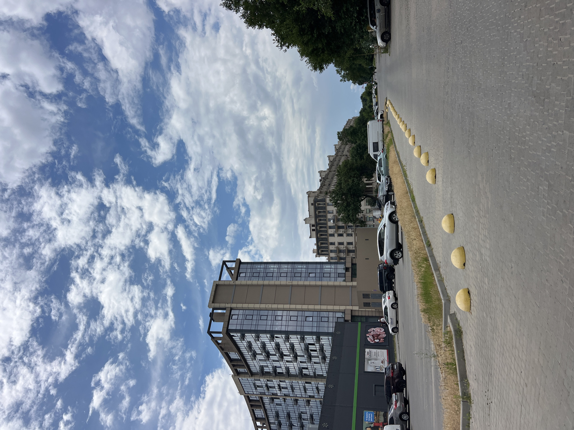 The image depicts a street scene in Fontanka, Ukraine, under a cloudy sky. In the foreground, a paved road with yellow hemispherical bollards lines the right side. To the left, a parking lot is visible, with several cars parked, including a white sedan, black SUVs, and a silver SUV. Behind the parking lot, a modern, multi-story building with a glass facade and balconies stands tall. Further down the street, other buildings and mature trees line the roadside. A white van is parked in the distance, and several people are walking along the street. The overall impression is of a bright, urban environment on a partially overcast day.