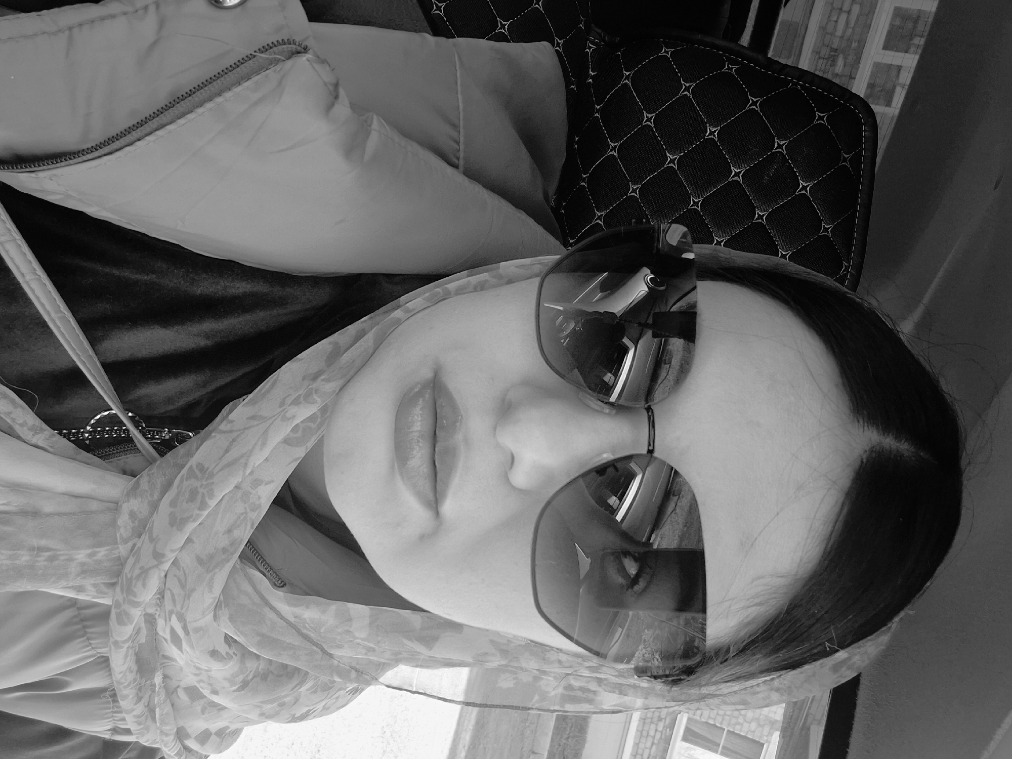 The black and white image, rotated 90 degrees counter-clockwise, features a person, likely a woman, in what appears to be a vehicle interior or similar setting in Chaikovsky, Russia. Her face is partially visible, dominated by large, dark, reflective sunglasses that obscure her eyes. Her dark hair is pulled back, and her full lips are slightly parted.
She is wearing a light-colored, possibly padded or puffy, jacket with a visible zipper on the front. A light-colored scarf, featuring a subtle floral or abstract pattern, is draped around her head and neck. To the upper right, a black surface with a distinctive quilted, diamond-stitched pattern is visible, suggestive of car seat upholstery or a bag. Reflections in the sunglasses show a blurred outdoor scene, including what appears to be a building with windows, indicating it is daytime.
The person appears to be posing for the camera in a calm manner. No specific activity is taking place beyond this pose. There is no visible text in the image. The overall impression is a casual portrait taken within a private, enclosed space with a view to the outside.
