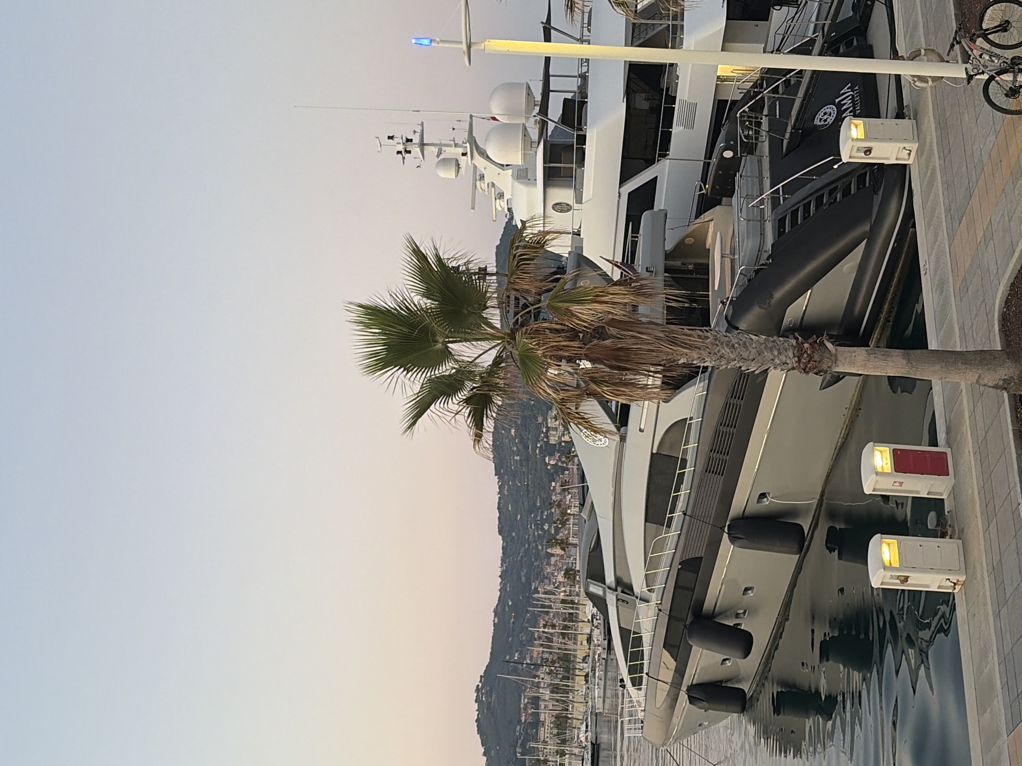 This media file captures a serene scene at a marina in Imperia, Italy, likely during the late afternoon or early evening. The primary subjects are large luxury yachts docked in the calm water, with a prominent palm tree in the foreground partially obscuring the view. The setting appears to be a waterfront promenade with a paved walkway.
The image showcases several large yachts, one of which is particularly detailed, showing its sleek, modern design with dark hull accents and white superstructure. Attached to its side is a large, black inflatable tender or dinghy. The yachts are moored alongside a concrete quay, where two white electrical hook-up boxes with illuminated lights are visible. One box has a red door, while the other has a white door.
In the background, a hillside town with numerous buildings and sailboats dotting the harbor creates a picturesque backdrop. The sky is a soft gradient of pale blue and warm, peachy tones, suggesting the approach of sunset. The weather appears calm and clear. A bicycle is leaning against the railing of the promenade, hinting at the presence of recreational activity.
There is visible text on the side of the large inflatable tender, which appears to be "DIANA VALLETTA" in white lettering. The overall mood is tranquil and peaceful, characteristic of a quiet harbor at dusk.