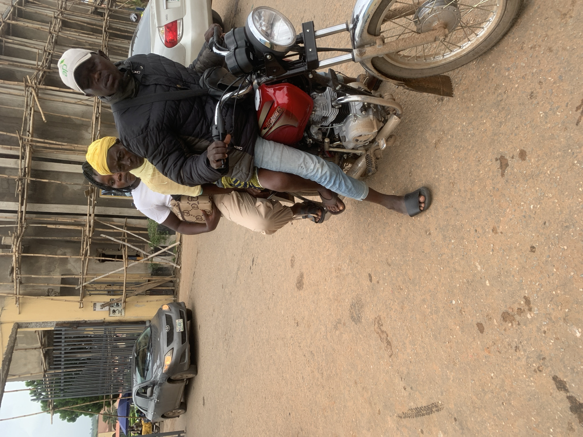 A group of three people, two women and one man, are riding on a motorcycle in Shagamu, Nigeria. The man, wearing a black jacket and a white cap with a green logo, is driving. A woman in a yellow headscarf and a woman in a white shirt are seated behind him. A gray car is parked to the left of the motorcycle. In the background, a building under construction with scaffolding made of bamboo is visible. The ground is a dirt road, and the overall scene appears to be a typical street in the city.