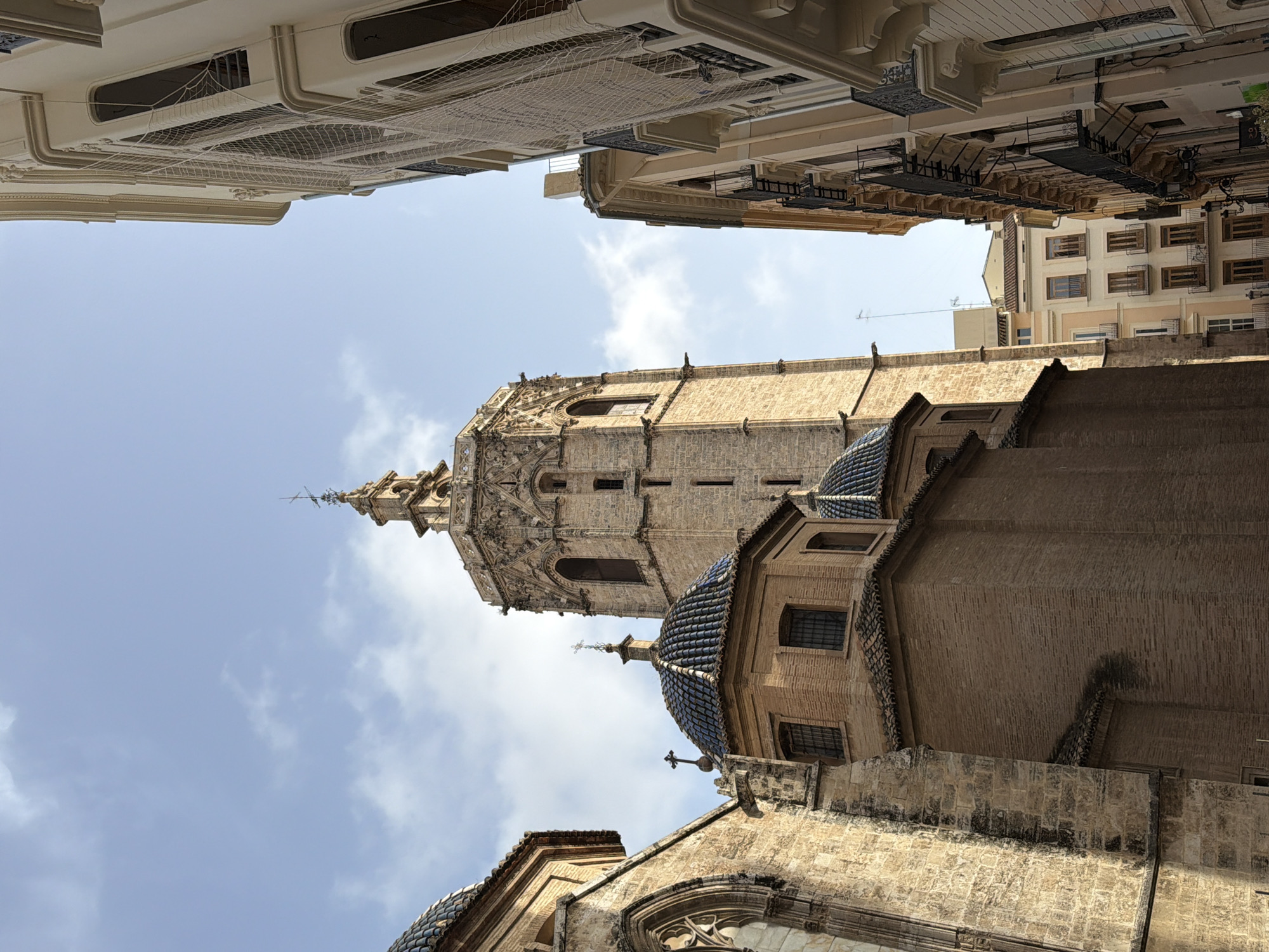 The image shows a view looking up at historic buildings against a sky with scattered clouds. In the foreground and to the left, a section of a large stone building with Gothic architectural elements and a blue-tiled dome is visible. Adjacent to this, a taller tower with intricate stone carvings and arched windows rises into the sky. To the right of the tower, a light-colored building with balconies and dark metal railings is shown, with a net covering its facade. Further back, several other buildings of varying heights and architectural styles are visible, some with tiled roofs. The overall scene depicts an urban environment with significant historical architecture.