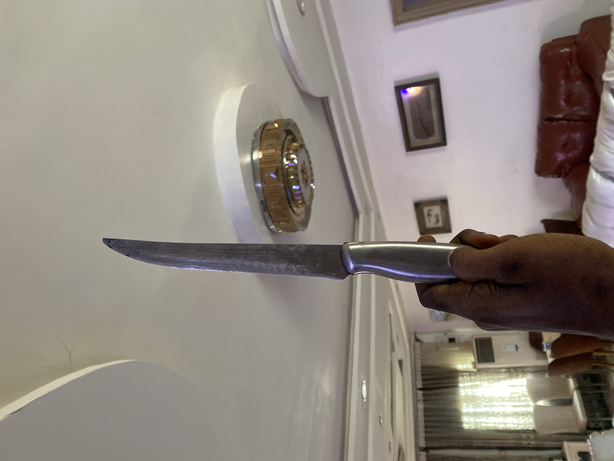 The image shows a person's hand holding a knife upwards, with the blade facing away from the viewer. The background appears to be a residential interior, possibly in Giginyu, Nigeria. The ceiling has ornate light fixtures and a textured design. There are framed pictures on the wall and furniture, including a red leather sofa, visible in the background. The lighting suggests it is daytime, with natural light coming through a window covered by curtains. The overall scene is mundane, with no immediate indication of violence or distress.
