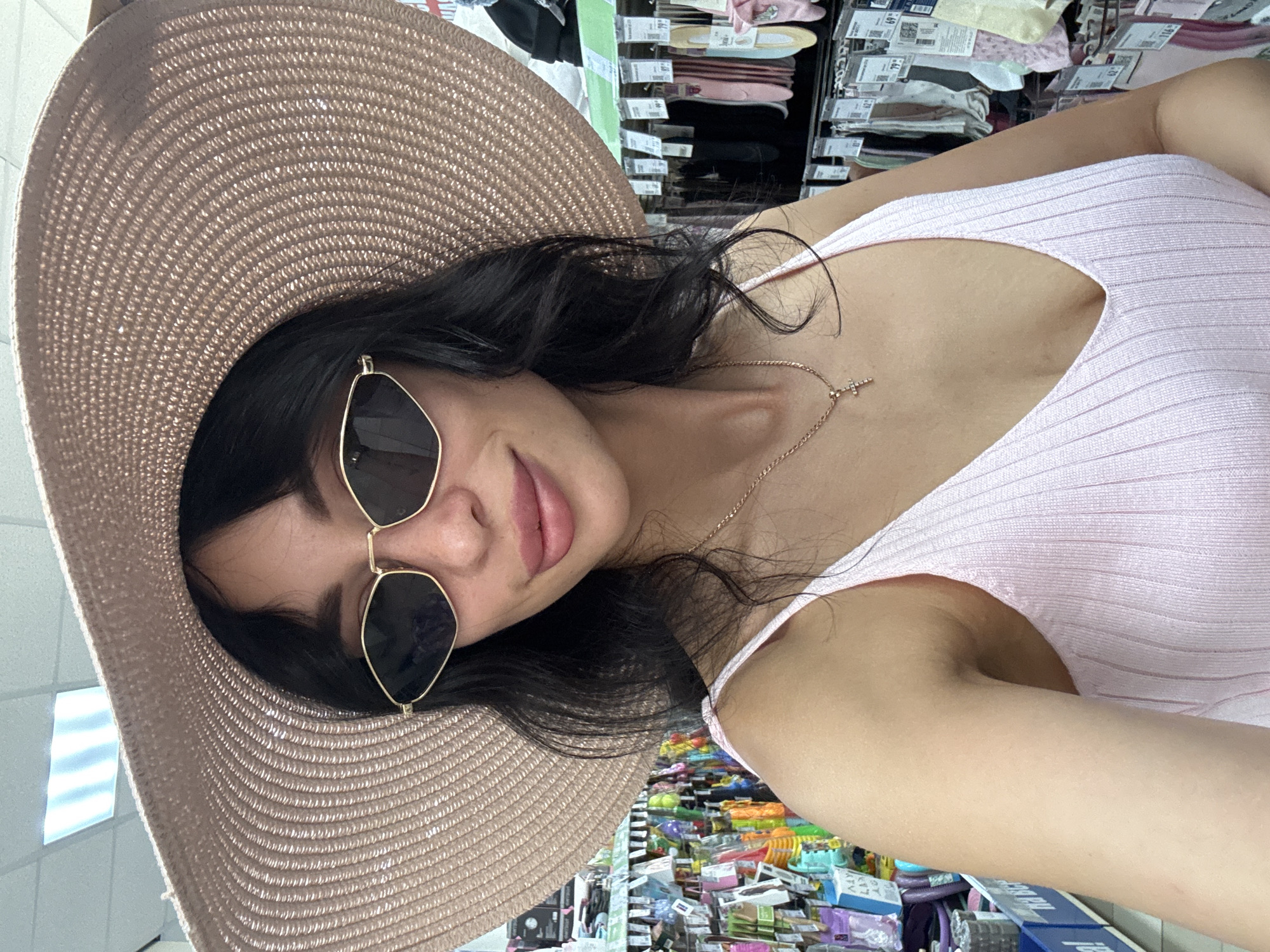 A medium shot shows a young woman in a pink tank top and a wide-brimmed straw hat, wearing sunglasses indoors. The background appears to be a retail environment, with shelves stocked with various products, possibly in a store located in Chaikovsky, Russia. The woman is looking at the camera, and her right arm is partially visible.