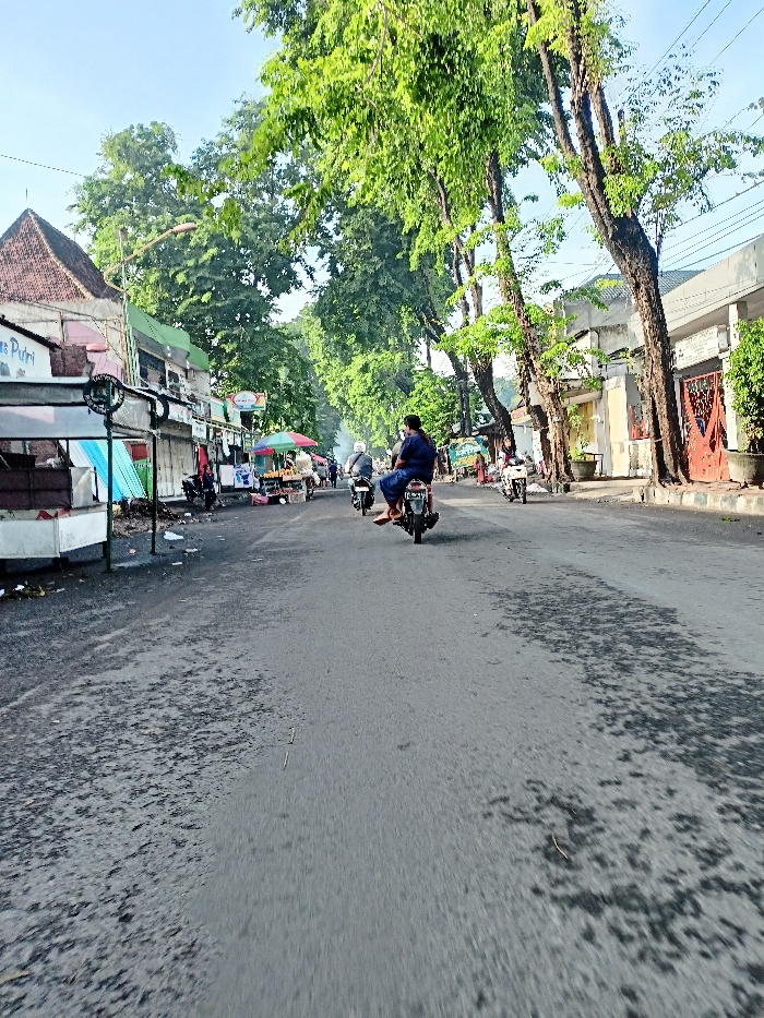 The image depicts a bustling street scene in Mojosari, Indonesia, likely during the early morning or late afternoon due to the clear sky and directional sunlight. The setting is an urban or semi-urban environment characterized by a paved asphalt road, numerous trees, and buildings lining both sides.
Several people are visible, primarily riding motorcycles. In the foreground, a person clad in blue clothing is seen from behind, riding a motorcycle away from the viewer. Further down the street, other individuals are also on motorcycles. On the left side, people are present near the vendor stalls.
The left side of the street features multiple vendor stalls or small shops, some with colorful umbrellas and canopies. One stall prominently displays fruits, while another, partially obscured, shows the text "Es Putri". Buildings with varying architectural styles, including one with a distinctive pyramid-like roof, stand behind these stalls. The right side of the street is lined with more permanent structures, possibly houses or shops, shaded by a row of tall trees with dense green foliage.
The activity captured is typical daily life, with people commuting on motorcycles and street vendors open for business. The worn asphalt road shows patches of darker moisture or stains. The overall impression is that of an active local street, characterized by vibrant greenery, commerce, and movement.