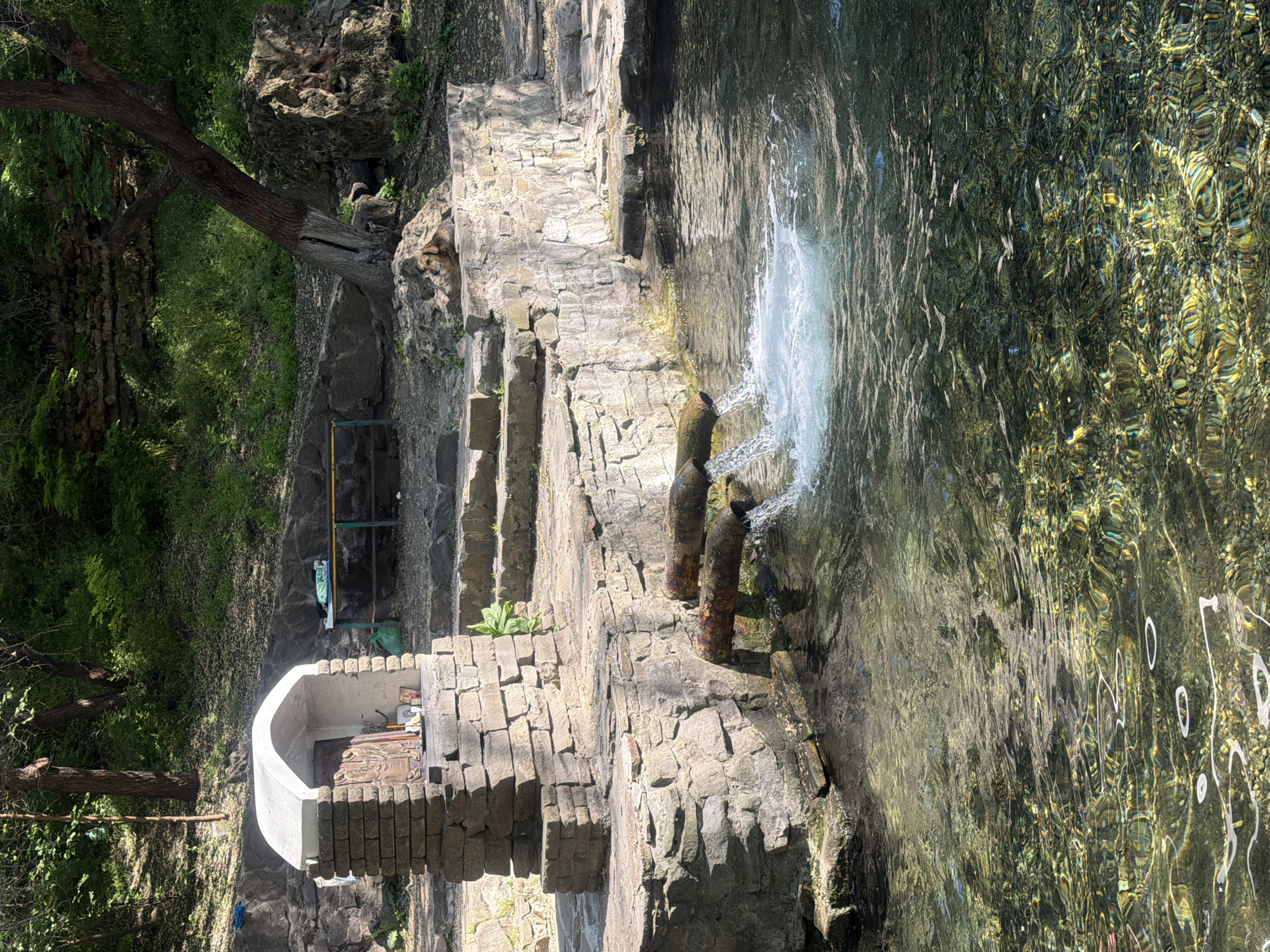 The image shows a water source and architectural structures in Rostov-on-Don, Russia.
A stone structure with an arched opening sits at the base of a hillside. Behind this structure, steps lead up the hill past a metal utility frame. Further up the hill, stone steps lead up to two rusty pipes from which water is flowing into a pool. The pool water is clear with light reflecting on its surface, creating a shimmering effect. Vegetation can be seen on the hillside. The scene is bathed in sunlight.