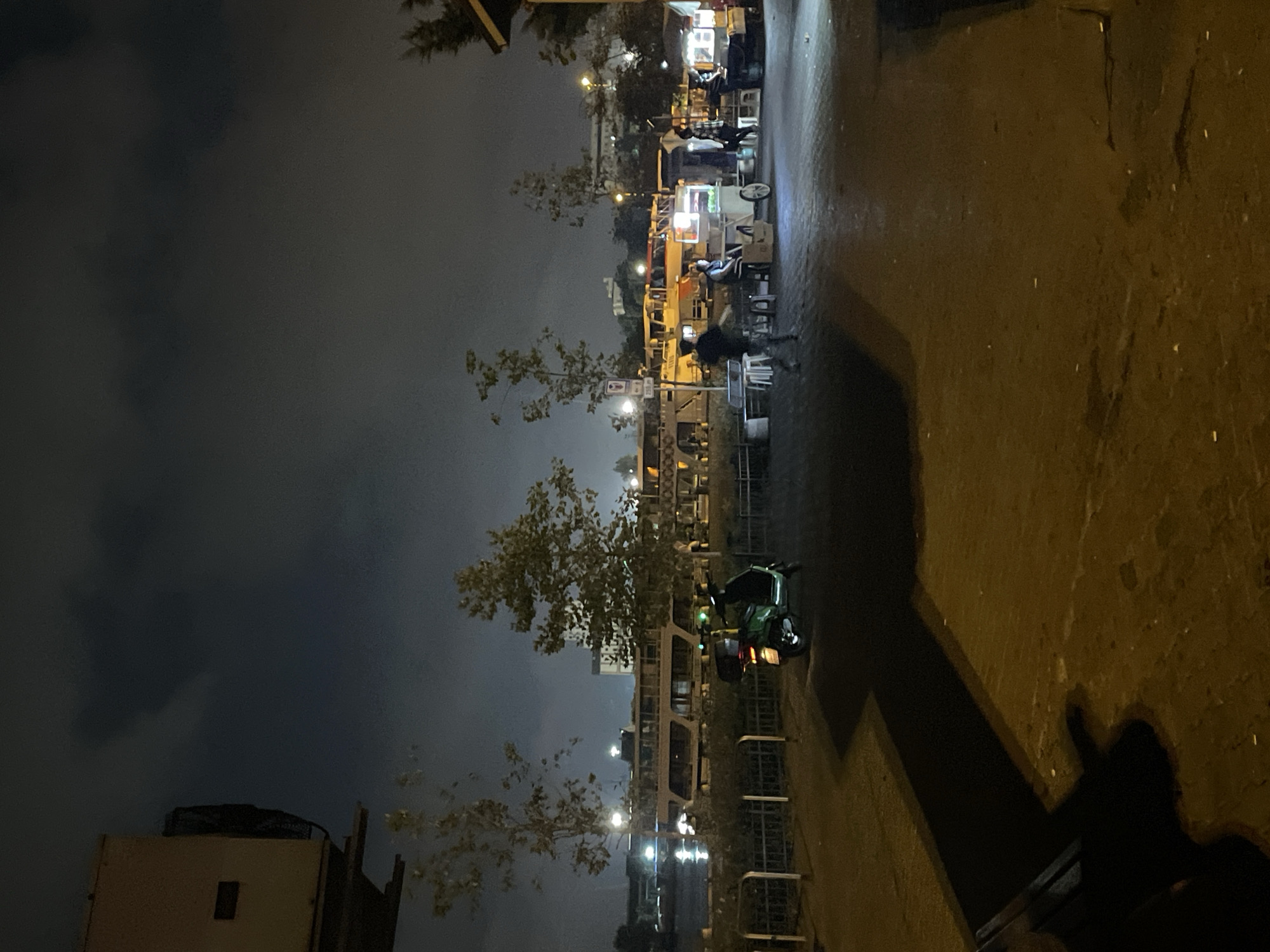 The image depicts a nighttime scene in Kadıköy, Turkey, likely near the waterfront. In the foreground, a paved area is illuminated by artificial lights, casting long shadows from unseen objects. A vibrant green scooter is parked on the left side of the paved area, facing right. Beyond the scooter, a metal fence runs horizontally, separating the paved area from a body of water where several boats and ferries are docked. The boats are illuminated by their own lights, reflecting on the water. In the mid-ground, several food vendors with carts and stalls are set up, some with people interacting around them. A few individuals are visible, some appearing to be customers and others vendors. The background features a cityscape with buildings and trees, all under a cloudy, dark sky. The overall atmosphere is that of a bustling nocturnal urban environment.