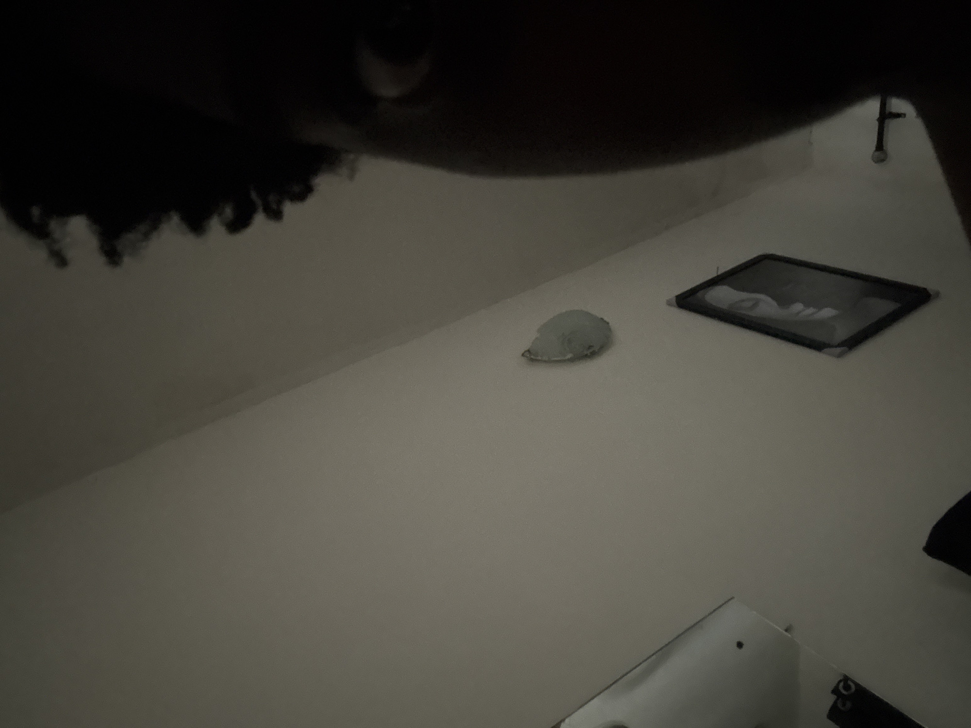 The image presents a dimly lit indoor scene from a low perspective, possibly looking across a surface. In the upper left quadrant, the top of a person's head with dark, curly hair is visible, suggesting the individual is leaning over or close to the camera.
The central part of the image features a light-colored, irregularly shaped object, which could be crumpled paper or a piece of light fabric, resting on a flat, light-toned surface. To its right, a dark-framed rectangular object, resembling a picture frame or a small tablet, lies horizontally. Inside this frame, a blurry, possibly monochrome image or reflection of a human face is discernible. Towards the upper right edge, a dark, vertical element, potentially a pipe or part of a fixture, is seen against a light-colored wall. In the bottom right corner, a reflective surface, possibly a mirror or another screen, is partially visible, alongside a dark object that seems to bear indistinct characters such as "GG" or "GO."
The low light suggests the time of day is likely evening or night. The setting appears to be a quiet room in Ihiagwa, Nigeria, with no specific activity taking place other than the static arrangement of objects. The overall mood is subdued due to the limited illumination.