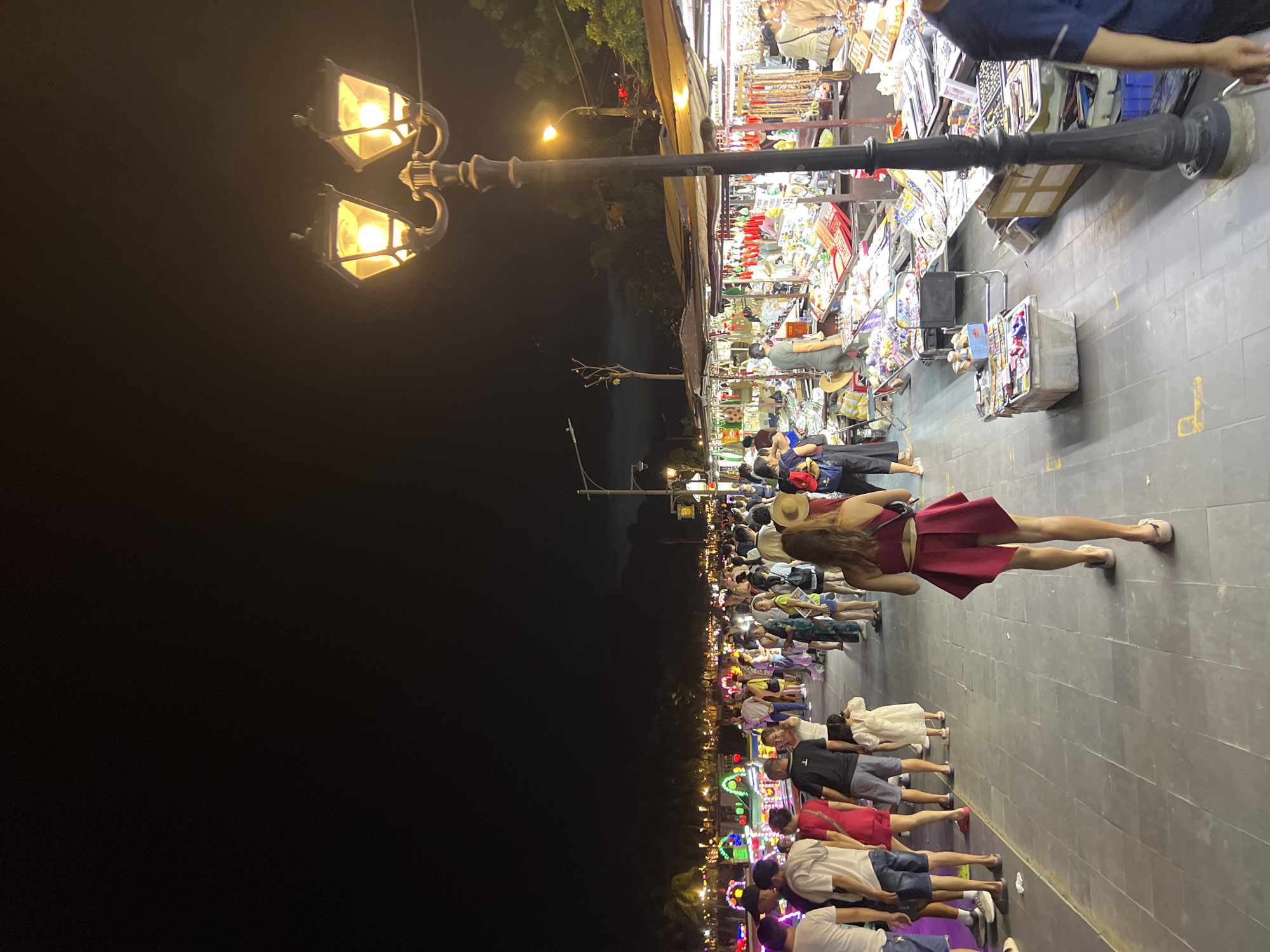 The image depicts a bustling night market scene in Hoi An, Vietnam, as indicated by the location context. The time of day is night, evident from the darkness and the illuminated streetlights and market stalls. The weather appears to be clear, allowing for outdoor activity.
The scene is a pedestrian walkway lined with numerous market stalls on one side and a dark, open area on the other, possibly a river or canal given Hoi An's geography. A tall, ornate black lamppost with two glowing, glass-enclosed lamps stands prominently in the foreground, casting a warm light.
A crowd of people is walking along the illuminated pathway, creating a sense of lively activity. In the foreground, a woman wearing a short, red dress and a straw hat is walking away from the camera, her posture suggesting leisure. Other individuals, dressed in casual attire, are visible in the background, some browsing at the stalls and others walking. The market stalls themselves are filled with various goods, likely souvenirs and local crafts, illuminated by colorful lights and bright bulbs.
The overall mood is one of energetic, nighttime commerce and recreation. The lighting creates a festive and slightly romantic atmosphere, common in night markets. No visible text is discernible in the image.