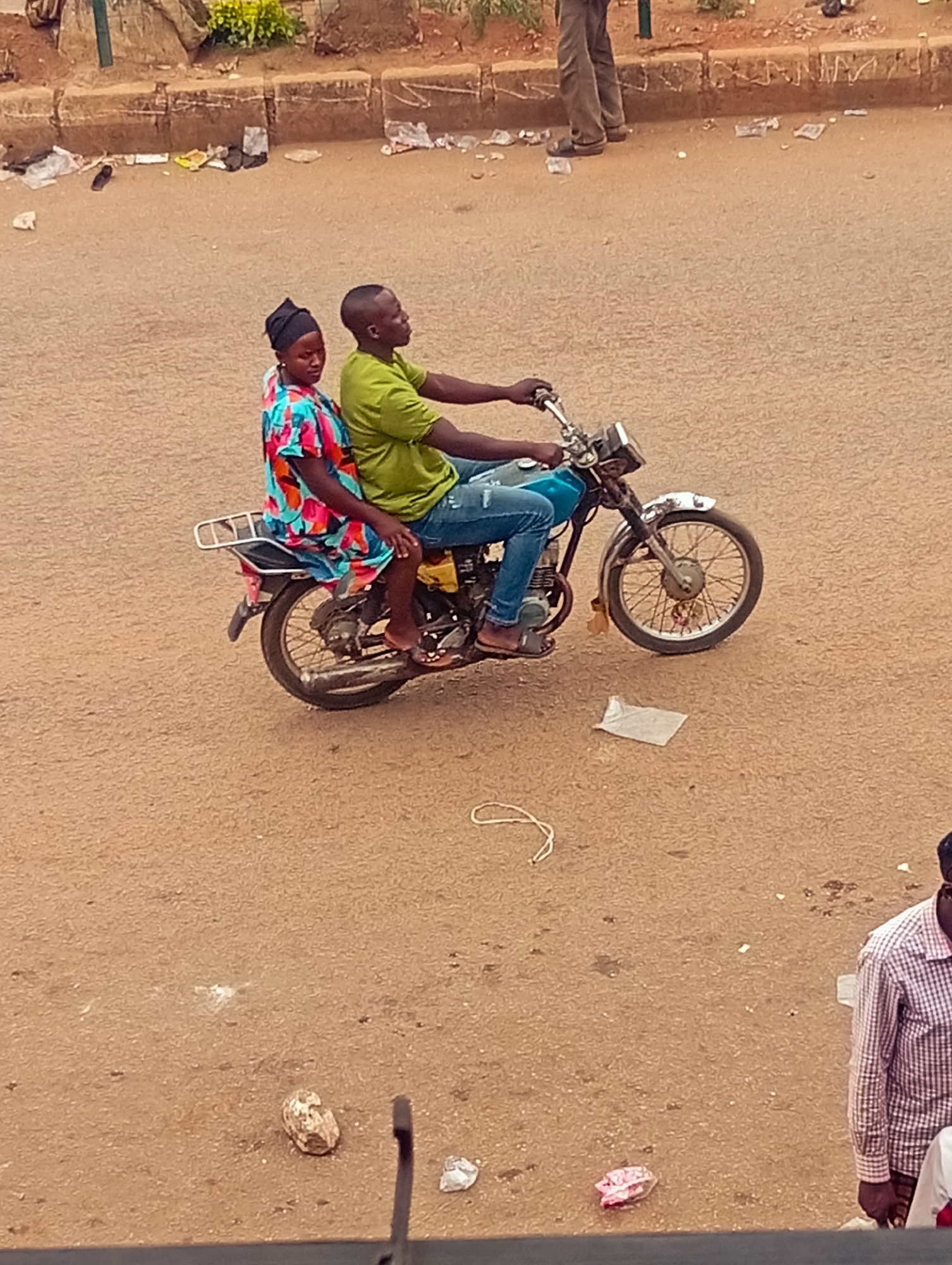 A man and woman are riding a motorcycle on a street in Unknown, Unknown. The man is driving, wearing a green shirt, blue jeans, and sandals. The woman is sitting behind him, wearing a colorful dress and a headscarf. The motorcycle is old and rusty. There is trash scattered on the street around them. Another man is standing on the right bottom corner of the image, wearing a plaid shirt.