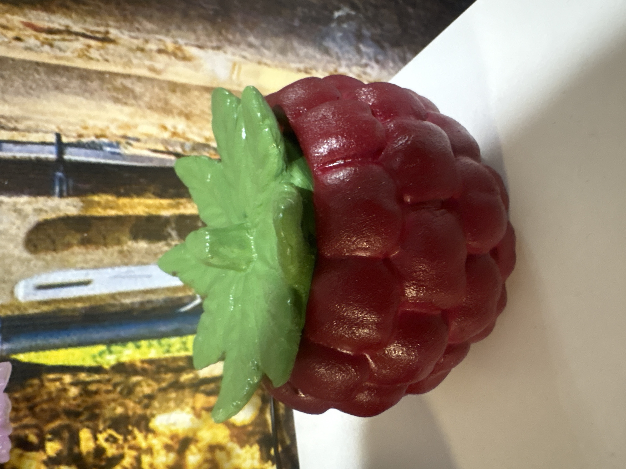 A raspberry-shaped, dark red container with a green leaf lid is centered on a white surface. The background consists of vertical, textured elements in brown and cream tones, with hints of yellow and blue. The lighting is from the front and slightly to the right, creating subtle shadows and highlights on the raspberry container. There are no people or animals visible in the image. The scene appears to be indoors, likely on a table or shelf.