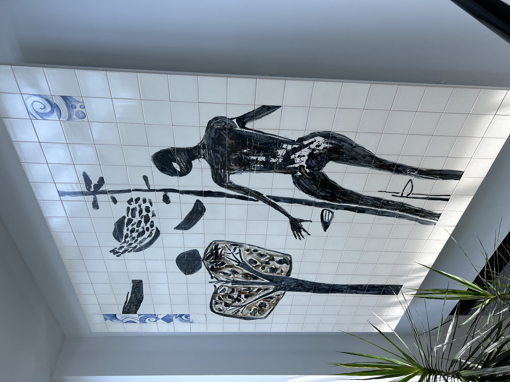 The image shows a large, tiled mural on a white wall. The mural features abstract figures and shapes painted in black and brown. On the left, a figure resembling a tree with dark brown foliage stands tall. To the right, a slender, dark figure with an elongated body and limbs is depicted. The figures are rendered in a stylized, almost silhouette-like manner, with some areas filled with solid black and others with brushstroke textures. Scattered around the main figures are smaller abstract shapes. The mural is bordered by white tiles, and in the upper corners, there are decorative tiles with blue patterns, one featuring a swirling design and another with a diamond pattern. In the foreground, a couple of leafy green plants with long, pointed leaves are visible. The lighting suggests an indoor environment, possibly a hallway or public space, with natural light casting shadows from the plants onto the wall.