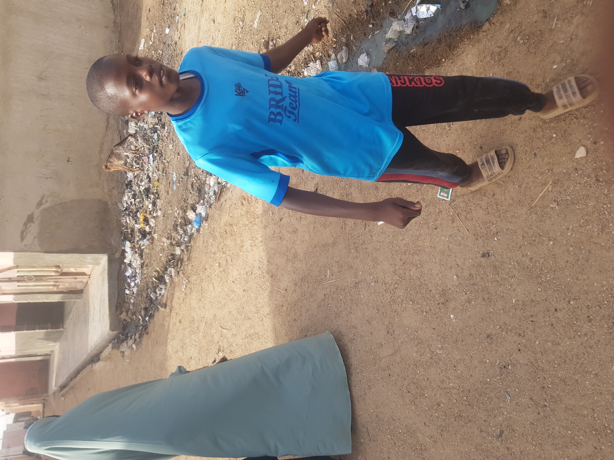 The image shows a boy standing on a dirt street in Dutsinma B, Nigeria. He is wearing a blue t-shirt with "Bride Team" printed on it, black pants, and sandals. There is trash visible in the background. A person wearing a light green robe is partially visible in the foreground. The street is unpaved and dusty. A building is visible to the left.