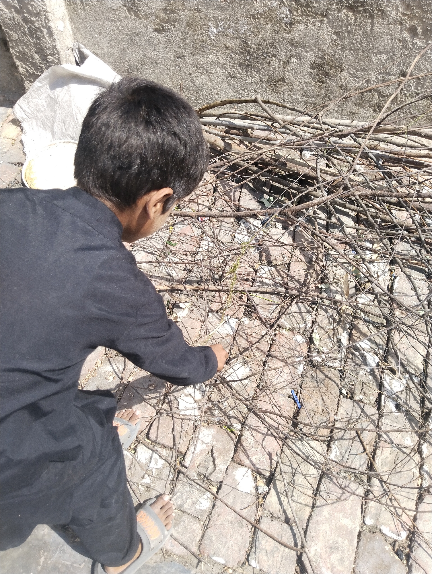 The image shows a child in Dera Chahal, Pakistan, wearing black clothing and sandals. The child is crouched near a pile of dry tree branches on a brick-paved surface next to a building wall. The child's right hand is extended towards the branches. A white sack is visible in the background.