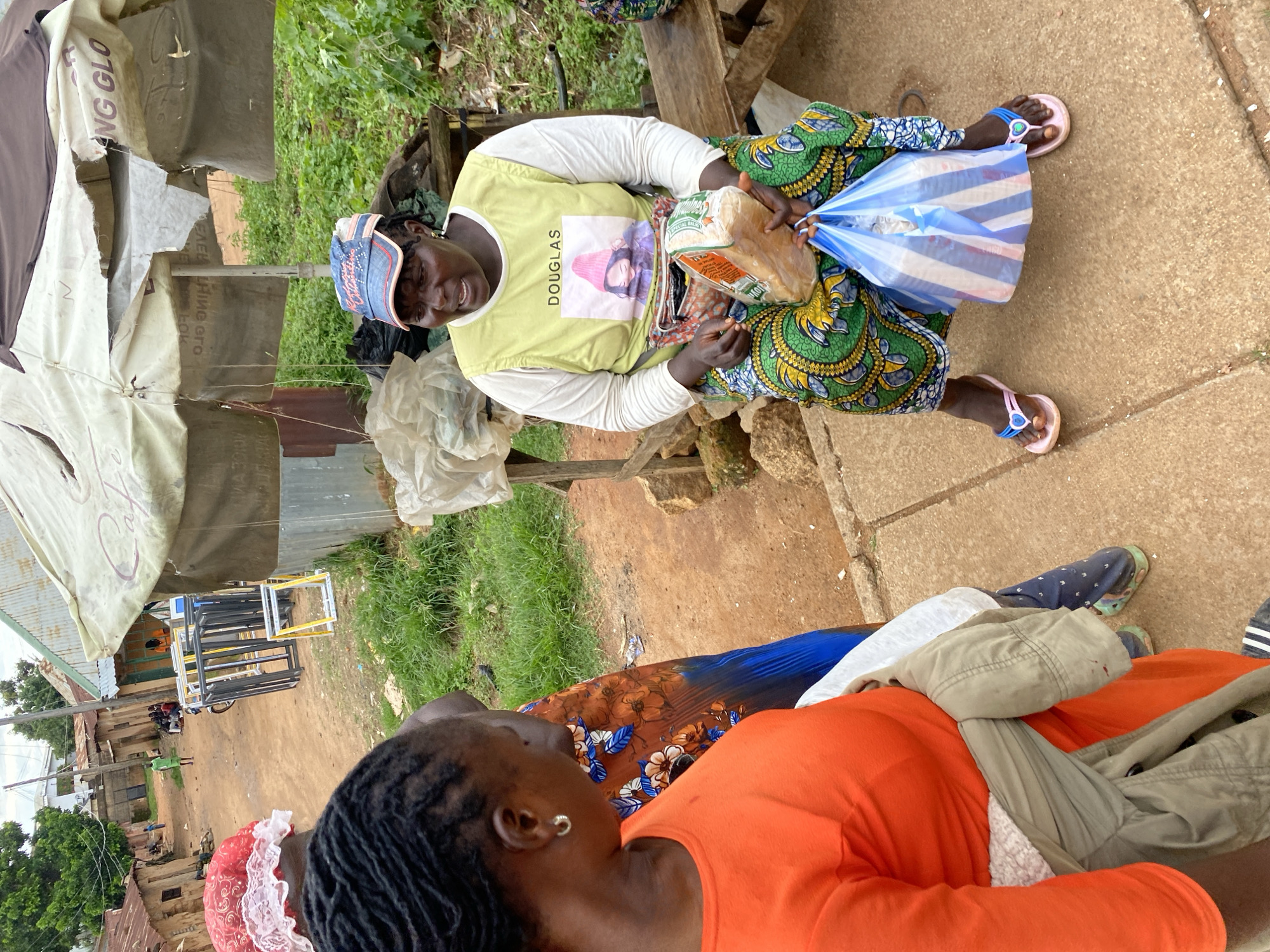 In Bukuru, Nigeria, a woman wearing a yellow vest with the name "DOUGLAS" and a printed image of a woman wearing a pink hat sits on a wooden bench. She is also wearing a blue and white patterned skirt, a red and white patterned headscarf, and blue and pink flip-flops. She is holding a loaf of bread wrapped in plastic in her right hand and a blue and white striped plastic bag in her left hand. To her left, another woman, seen from behind, wears an orange long-sleeved shirt and a grey jacket tied around her waist. The background shows a dirt road lined with buildings, green vegetation, and a market stall with a tattered umbrella providing shade. A motorcycle is parked on the road, and several other people are visible in the distance.