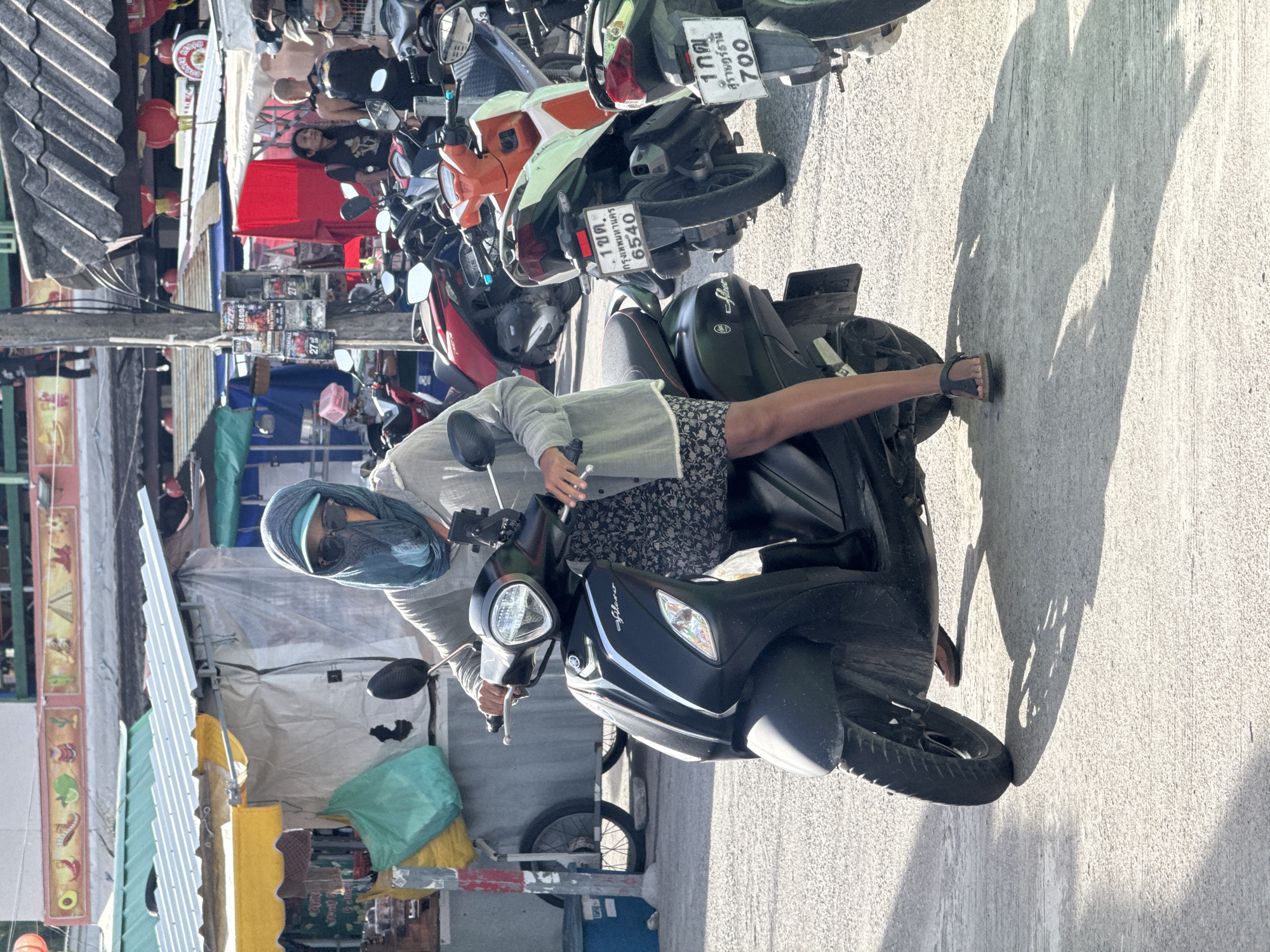 A person wearing a face covering and sunglasses is seated on a black Yamaha scooter, parked on a paved street in ตำบลเกาะพะงัน, Thailand. The person is dressed in a light gray long-sleeved shirt and a dark floral print skirt. They are wearing dark sandals. The scooter has a license plate that reads "1กฎ กรุงเทพมหานคร 6540". In the background, there are other parked scooters, market stalls with awnings, and people standing. The scene appears to be a busy street or market area.