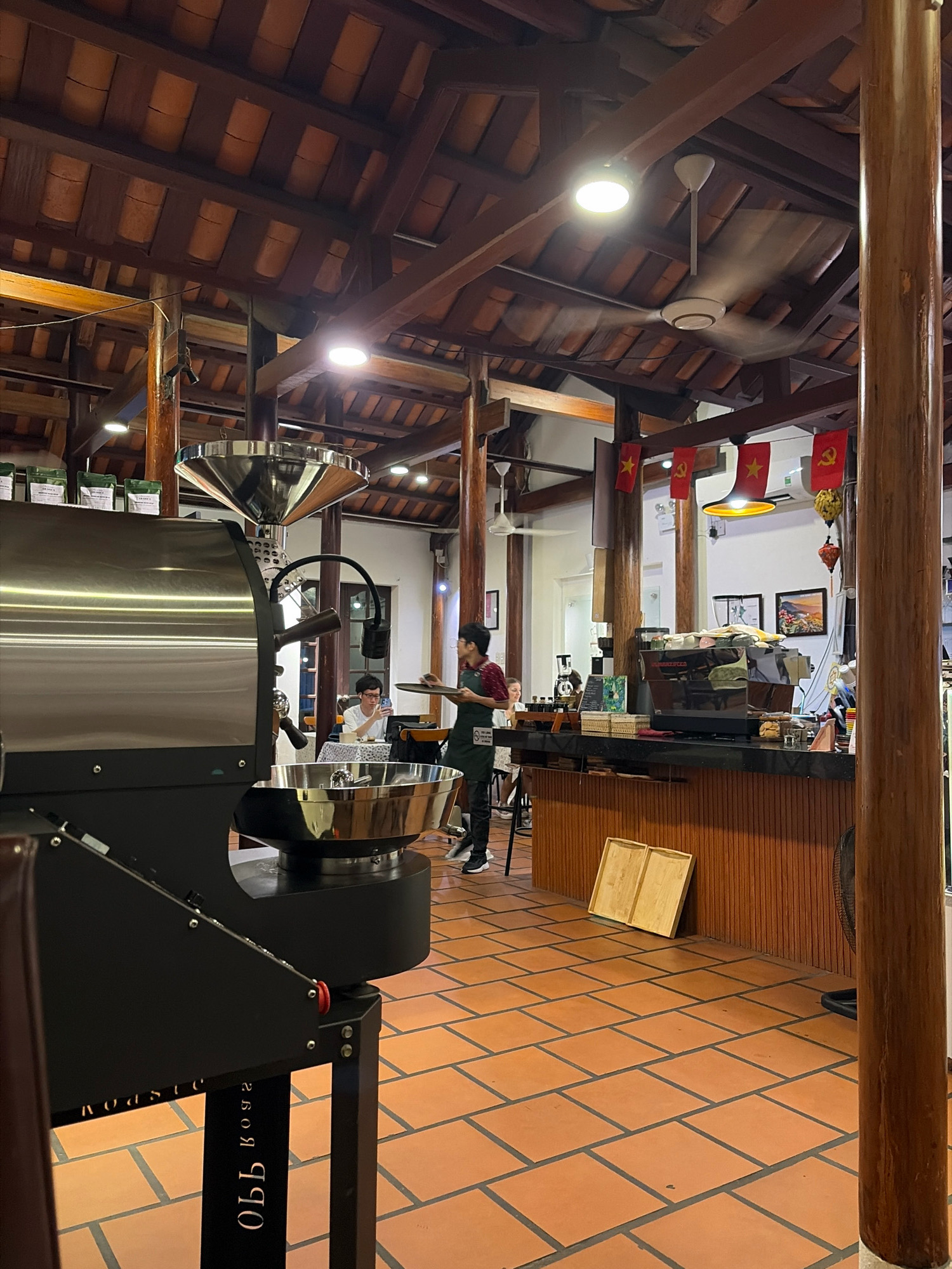 The image depicts the interior of a coffee shop with exposed wooden beams and a tiled ceiling. In the foreground, a large, stainless steel coffee roaster is prominent. The floor is tiled with orange terracotta squares.
In the mid-ground, a counter area features a professional espresso machine and various coffee-making equipment. Behind the counter, a person wearing an apron is visible, possibly a barista. A customer sits at a table to the left, looking at their phone.
The background shows a ceiling fan and lighting fixtures. Red flags with yellow stars are hanging from the ceiling, suggesting a political or nationalistic theme. There are also framed pictures on the wall. The overall atmosphere suggests a functioning coffee shop, likely in Đà Nẵng, Vietnam, given the context.