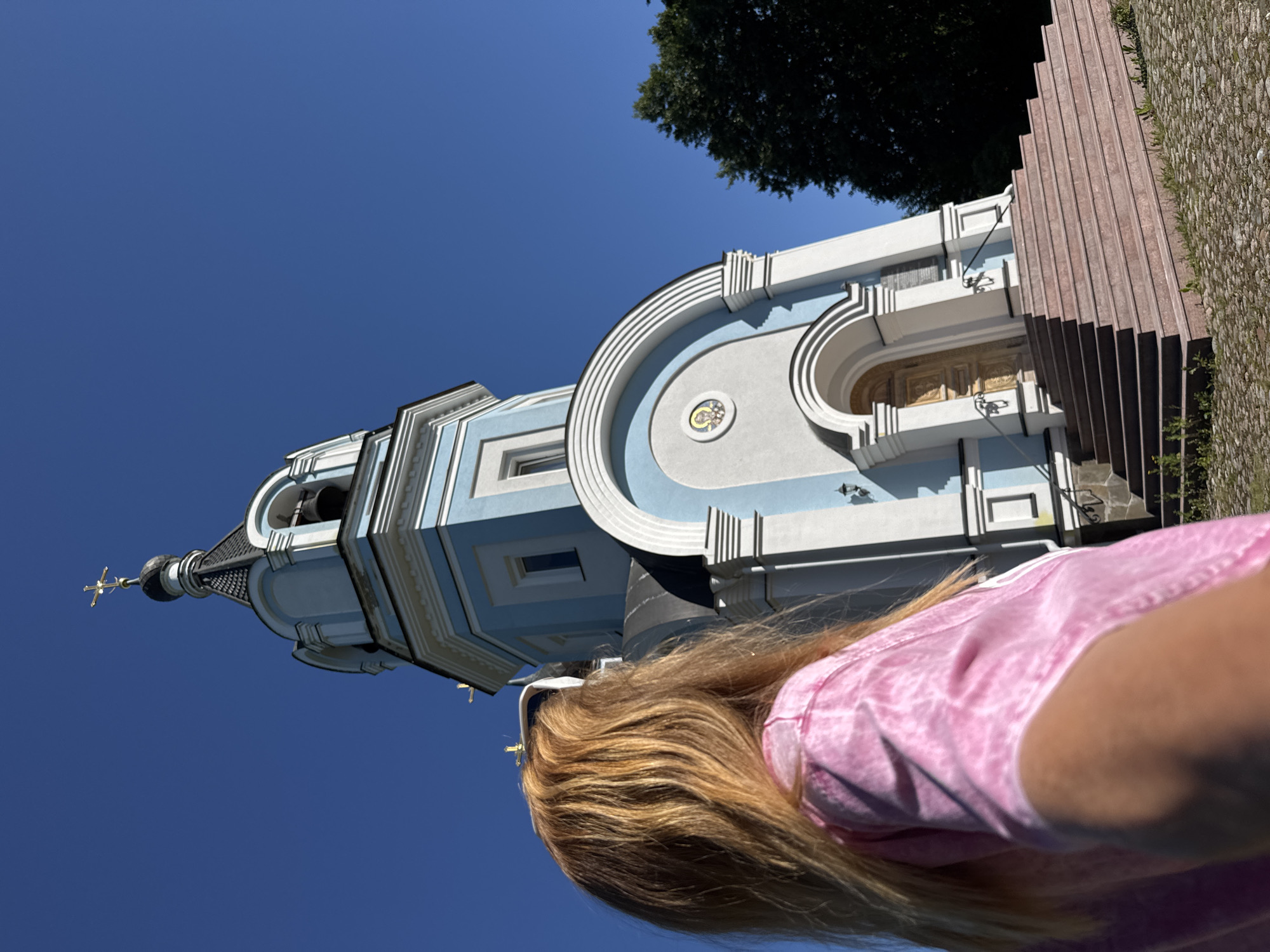 A person with long blonde hair, wearing a pink tie-dye t-shirt, stands facing away from the camera, looking up at a pale blue and white church. The church has a tall bell tower topped with a golden cross and a dark roof. A bell is visible in the tower. The church entrance features a large arched doorway with ornate wooden doors, accessed by a wide, multi-tiered staircase. The building is decorated with white trim and architectural details. A circular mosaic icon is set above the entrance. The scene is set against a clear blue sky, with a dark green tree visible to the right of the church. The ground in front of the church is paved with cobblestones and scattered with sparse green vegetation. The image appears to be taken from a low angle, emphasizing the height of the church. The location is St. Ozerki, Russia.