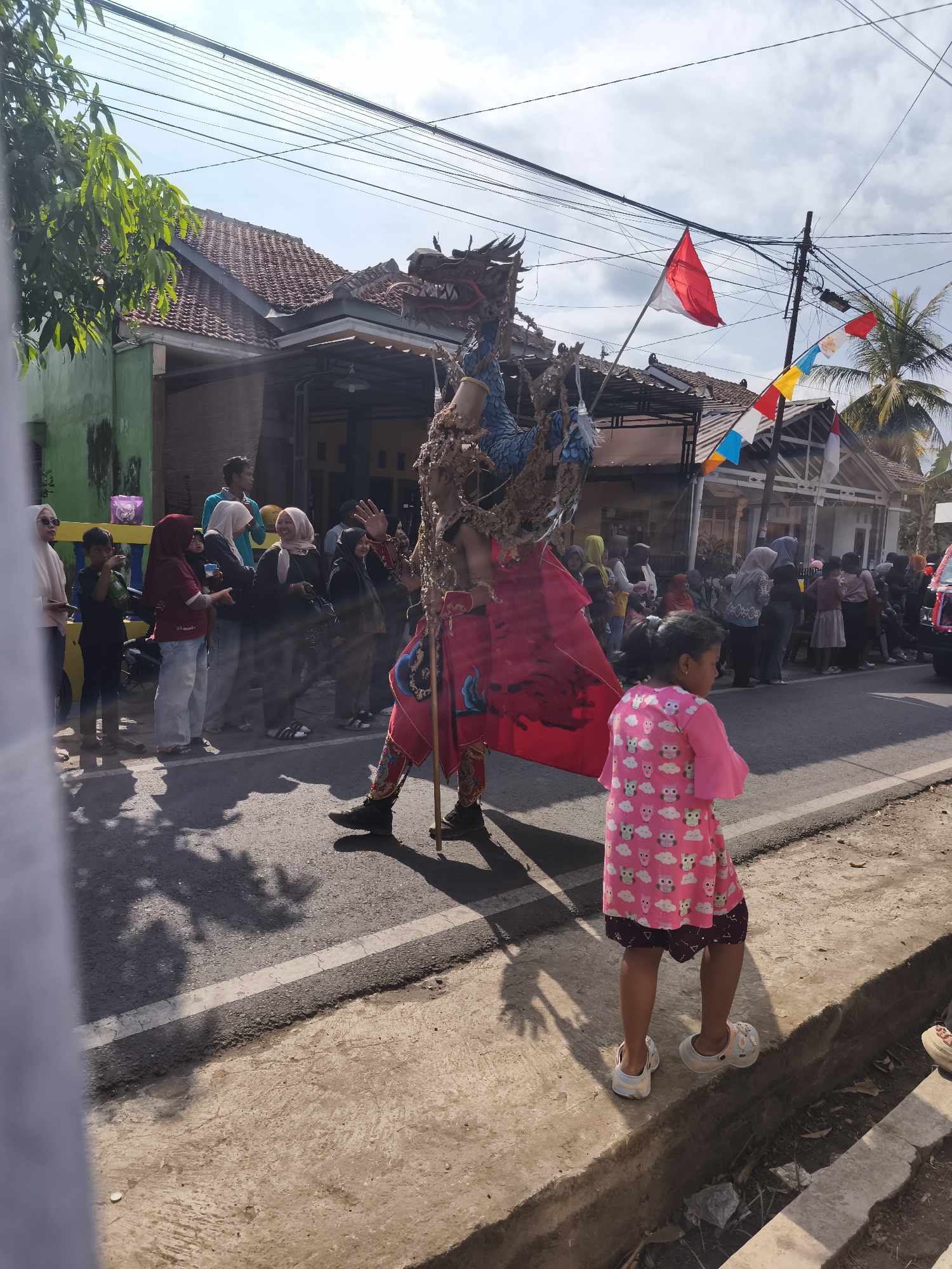 In Kutoarjo, Indonesia, a street scene unfolds under a bright sky with scattered clouds. A person in a vibrant, elaborate costume, designed to resemble a dragon with blue and red elements, stands in the middle of the road, holding a pole. The costume features a large red cape adorned with golden designs. The individual appears to be performing or participating in a parade, as indicated by their attire and the presence of onlookers.
To the right of the costumed figure, a young girl in a pink dress decorated with owl and cloud patterns stands on the side of the road, facing away from the camera. She is wearing light-colored clogs. Spectators, including adults and children, are gathered on the sidewalks and along the street, observing the spectacle. Many are wearing headscarves and traditional Indonesian clothing.
The environment suggests a residential area, with houses and tiled roofs visible in the background. Indonesian flags, red and white, along with colorful triangular banners, are displayed, suggesting a festive occasion or national celebration. Power lines crisscross overhead, a common sight in Indonesian urban and suburban landscapes. The road is asphalt with a white dividing line. The sun casts distinct shadows of people and trees onto the road, indicating it is daytime. The overall impression is of a lively community event taking place on a sunny day.