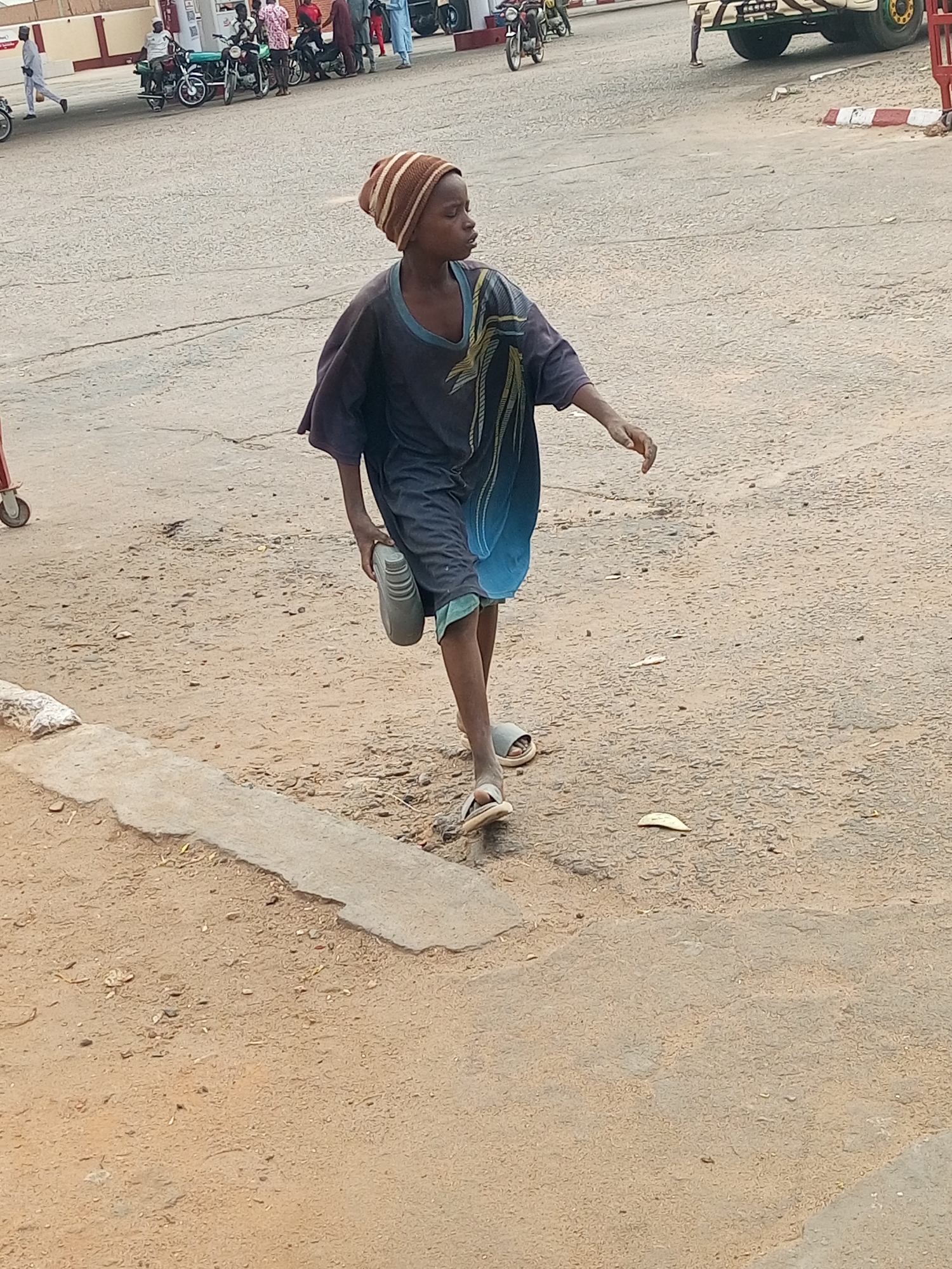 A young person wearing a striped beanie, dark clothing, and sandals is walking in an outdoor location. They are carrying a grey object, possibly a shoe, in one hand. The ground is a mix of concrete and dirt. In the background are motorbikes, a building with a red and white stripe, and a vehicle. Based on the style of the clothing and the presence of motorbikes, the location is likely Tempure, Nigeria.