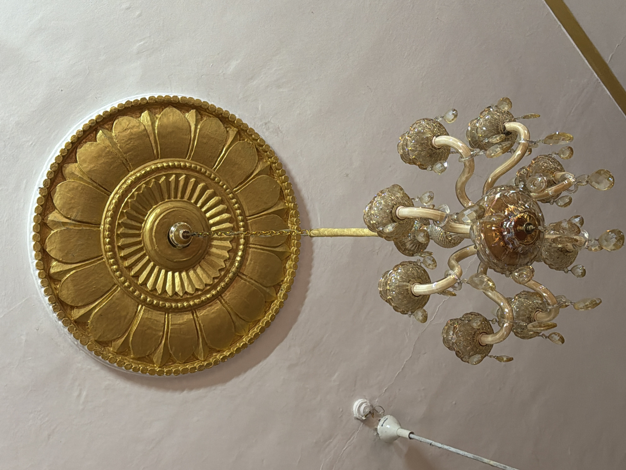 A close-up, overhead shot captures a section of a ceiling with ornate decorative elements and a chandelier. Dominating the left side of the frame is a large, gold-colored ceiling medallion with intricate floral patterns. The medallion is circular, with layered petals radiating outwards from a central, ribbed circular design. A delicate gold chain hangs from the center of the medallion, connecting to the chandelier.
To the right, a multi-armed chandelier hangs from the ceiling. The chandelier is made of a metallic material, likely gold-toned, and features several arms that curve outwards. Each arm holds a decorative glass element that appears to be a lamp shade or a decorative fixture, resembling clustered glass flowers or bulbs. Small, faceted glass crystals are suspended from various points on the chandelier, catching the light and adding a sparkling effect. The chandelier's central body is also a detailed glass or metallic element.
The ceiling itself is a light, neutral color, possibly white or a pale pink, with a textured surface that suggests plaster. In the lower right quadrant of the image, a small, white electrical fixture with a metal rod extending from it is visible, possibly an antenna or part of a fan, attached to the ceiling. The lighting in the scene appears to be a mix of ambient light, possibly from an unseen window or overhead source, and the subtle glow from the chandelier itself, though the lights on the chandelier are not illuminated. The overall impression is one of a decorative interior space, likely in a home or a building with traditional or opulent design elements, consistent with a location context in New Delhi, India. No people are visible in the frame. There is no visible text in the image.