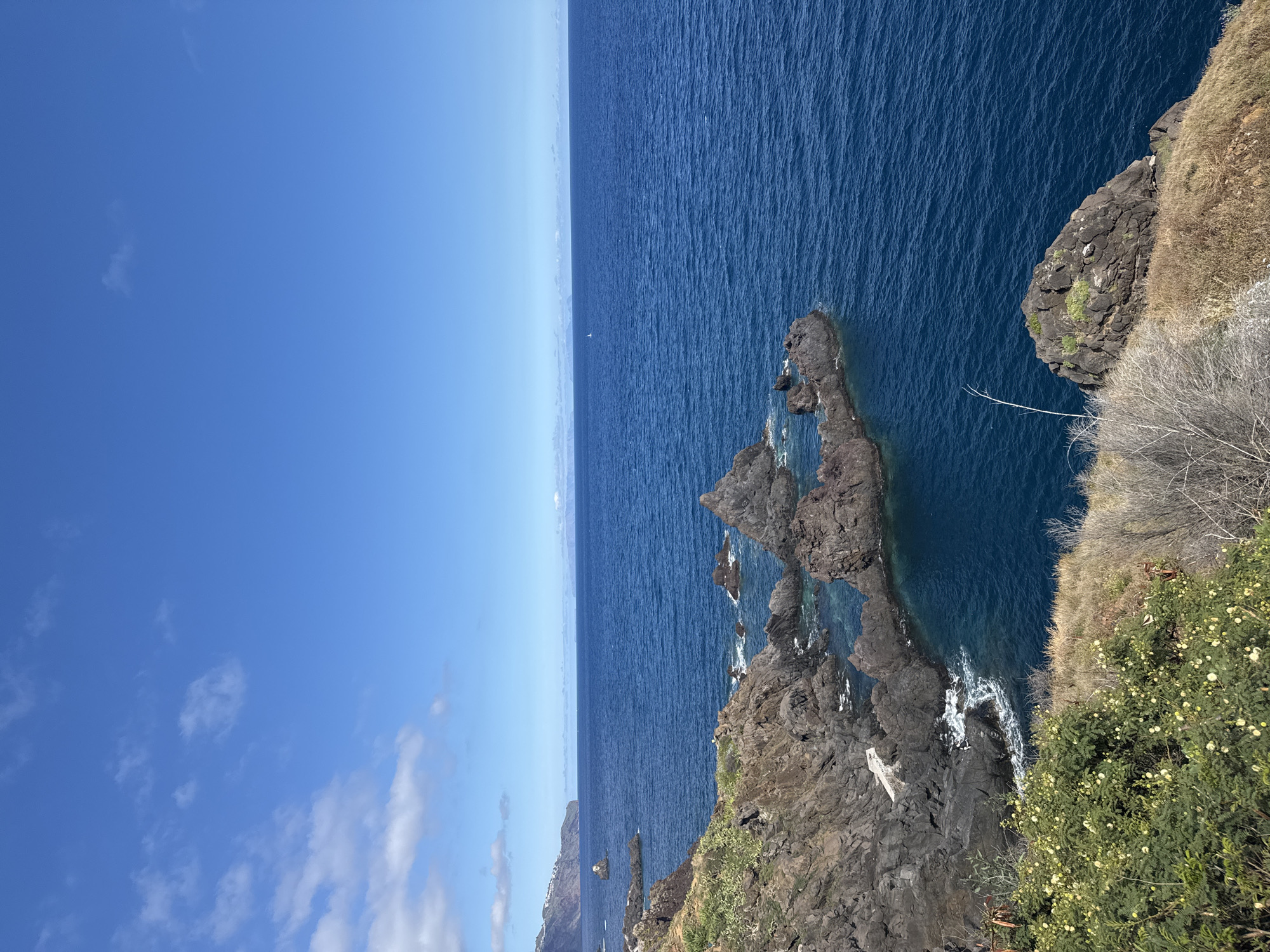 A long shot depicts a scene from Funchal, Portugal, featuring a cliffside view overlooking the ocean. The sky is blue, partly cloudy, with the horizon line visible in the middle of the frame. The ocean occupies the right half of the image, exhibiting deep blue water with some small waves. The cliffside on the right is covered in rocks, vegetation, and brown grasses. Some rocky outcroppings jut into the ocean with the ocean water lapping around them. A small sailboat is seen on the horizon.