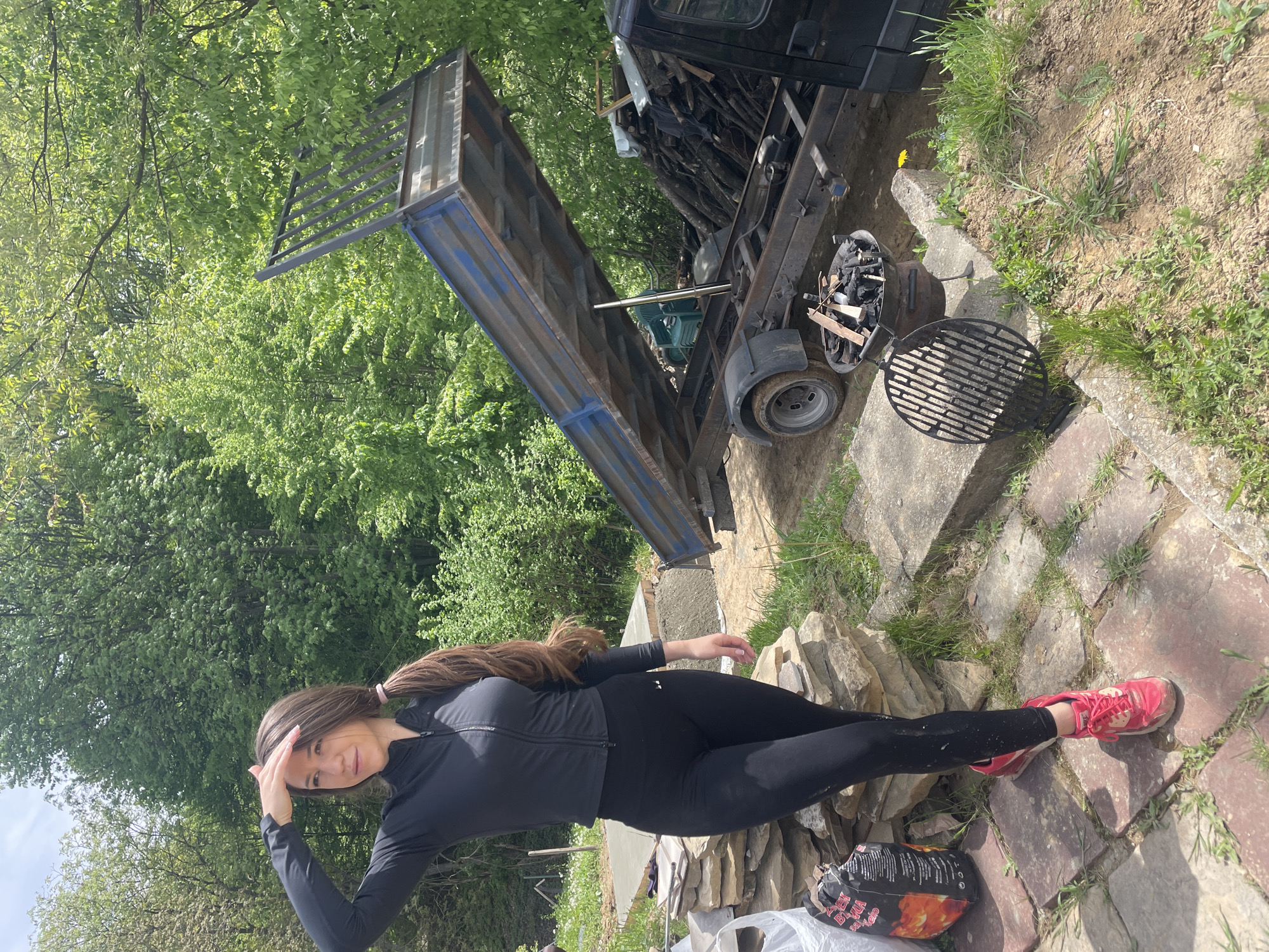 The image shows a woman in black athletic wear standing outside near a small utility trailer with a raised bed. She is holding her hand to her forehead, possibly shielding her eyes from the sun. The environment includes greenery suggesting a rural or suburban setting, and a barbecue grill is visible near the trailer. A bag of charcoal is near the feet of the woman on a stone paver. The precise location is Bogdanchovtsi, Bulgaria.