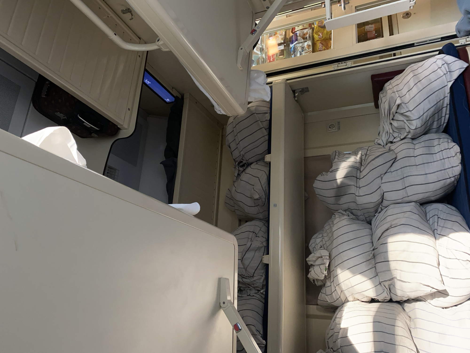The image depicts the interior of a compartment, likely a train carriage, with multiple berths stacked vertically. The berths are covered with bedding that appears to be in bags, all featuring a striped pattern of thin black and white lines. The lower berths are densely packed with these bagged items, creating a bulky, almost mountainous appearance. The upper berths also contain similar bagged bedding, with one visible suitcase placed on a shelf above. The overall environment suggests a travel context, possibly a long-distance train journey. The image does not provide any direct information about the city of Dryazgi, Russia, or any people present.