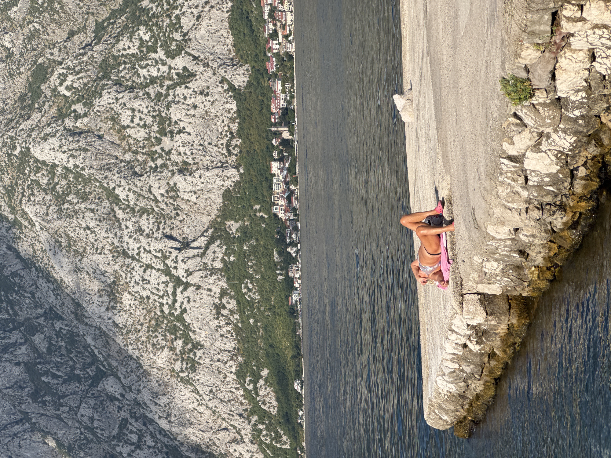 A woman is sunbathing on a concrete pier in front of a body of water. She is lying on her stomach on a pink towel, wearing a bikini. Behind her, the water extends to a shoreline with a town nestled at the base of a large, rocky mountain. The town consists of white buildings with red roofs and a church steeple. The mountain is covered in sparse vegetation. The scene is lit by sunlight.