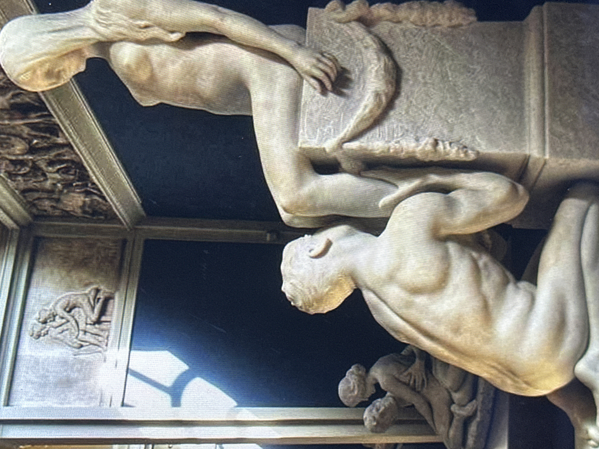 The image shows a sculpture of two figures in a classical style. In the foreground, a male figure kneels and appears to be interacting with the legs of a seated female figure. The female figure is partially draped, with a hood or veil covering her head and shoulders. Behind them, to the left, there is another sculptural relief depicting two figures embracing. To the upper right, there is a detailed frieze with multiple figures. The sculptures are light in color, likely marble or a similar material, and are set against a dark background. The lighting casts shadows on the figures and the surrounding environment. The location is described as Unknown, Unknown.