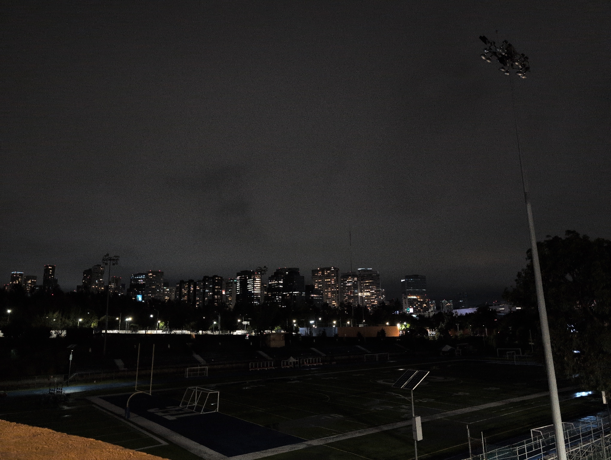 The image captures a nighttime view of a sports field, possibly a football or soccer stadium, within a city. In the foreground, a section of a dirt mound is visible on the left, suggesting a viewing area or embankment. The sports field itself is dark, with green turf marked with white lines. Two sets of goalposts are present on the field. A light pole with several lamps illuminates a portion of the field, casting long shadows. Further back, bleachers can be seen, also appearing dark and empty. Beyond the stadium, a dense line of trees separates the field from a cityscape dominated by tall buildings. Many of these buildings are illuminated, showing windows and architectural details against the dark sky. A tall, thin light pole extends from the right side of the image upwards towards a cluster of lights at its apex. The sky is dark and overcast, with the lights from the city reflecting faintly on the low-lying clouds. The overall impression is an urban scene at night, with a sports facility in the foreground and a bustling city skyline in the background. The location is identified as Mexico City, Mexico.