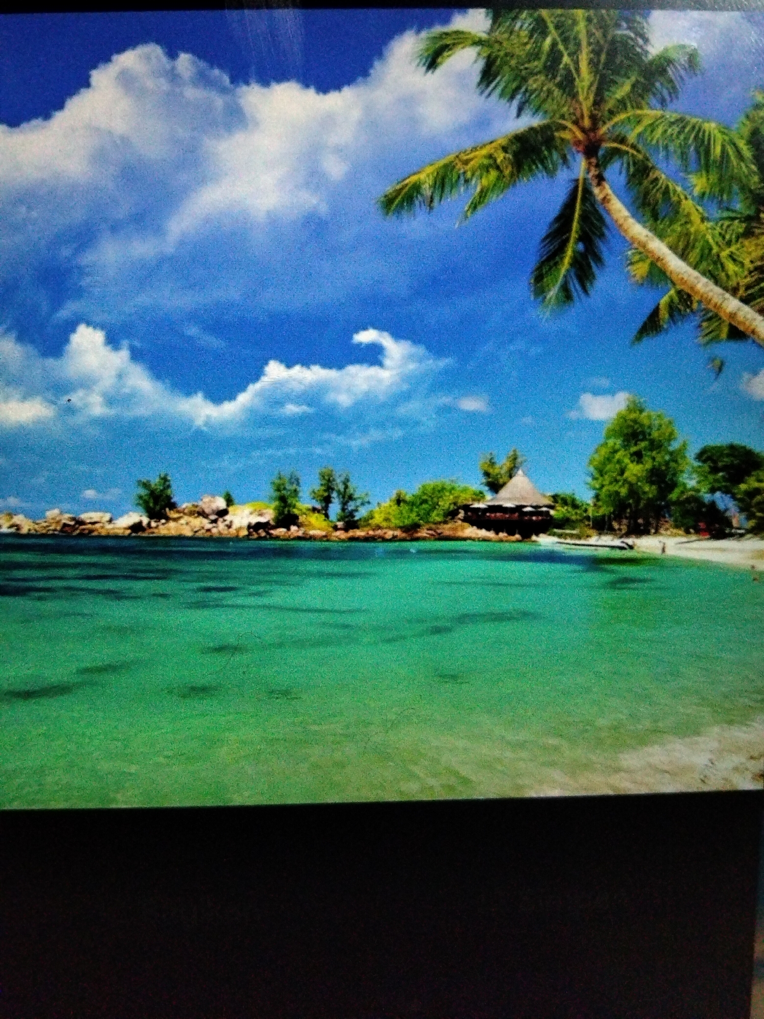 The image depicts a tropical beach scene with a clear turquoise ocean in the foreground. In the mid-ground, a sandy beach is lined with lush green trees and some rocky outcrops. A thatched-roof building, possibly a bungalow or restaurant, is visible among the trees. The background features a bright blue sky with scattered white clouds and a palm tree in the upper right corner. The scene appears serene and idyllic, suggesting a vacation destination. There are no visible people or specific landmarks to suggest a location within Kualaraja, Indonesia, or any other city.