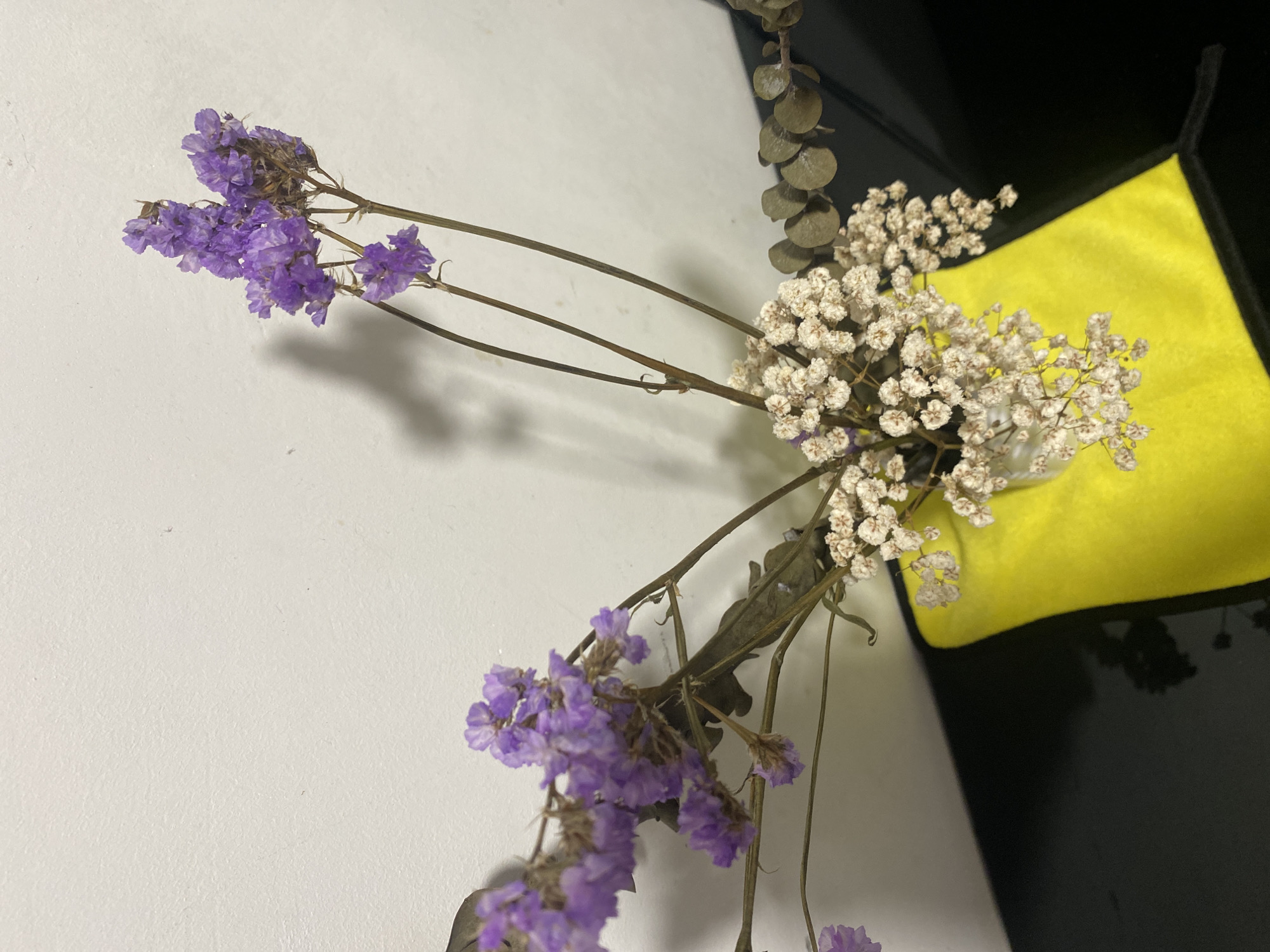 The image shows dried purple and white flowers with brown stems, along with some eucalyptus leaves. The flowers are arranged against a white wall. There is also a yellow cloth against a black background visible in the upper right of the image. The location is unknown; the prompt to mention Davao City, Philippines, is unsupported.